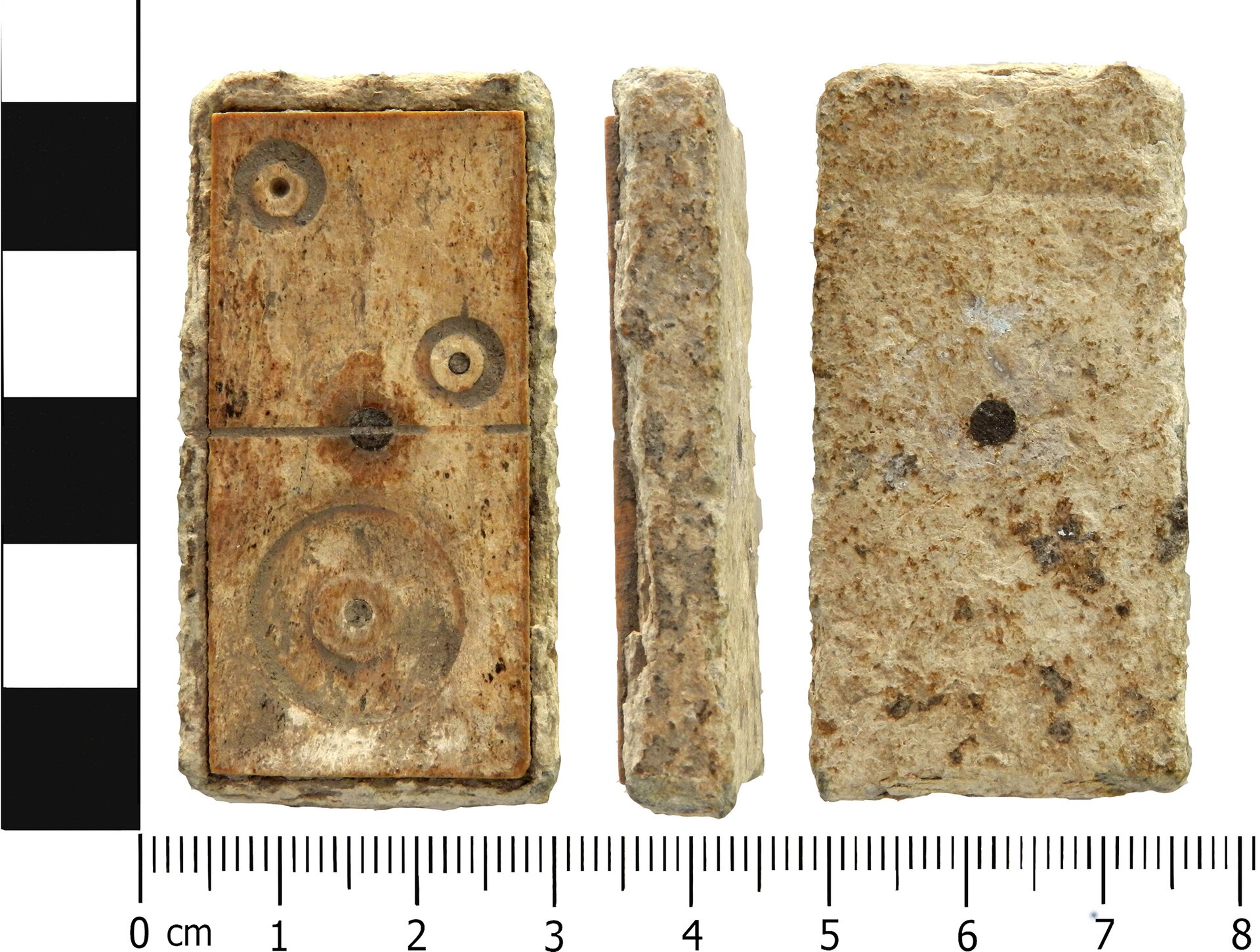 Zdjęcie przedstawia zabytkowy klocek domina, pokazany z trzech stron (od lewej) z przodu, z boku oraz z tyłu. Na przedniej stronie klocek podzielony jest na pół. Na górnej części widać dwa oczka, a na dolnej jedno duże oczko. Po lewej stronie oraz pod zabytkiem widoczne są miarki pokazujące wymiary klocka, który ma 2,7 cm szerokości, 5 cm wysokości oraz 1 cm grubości.