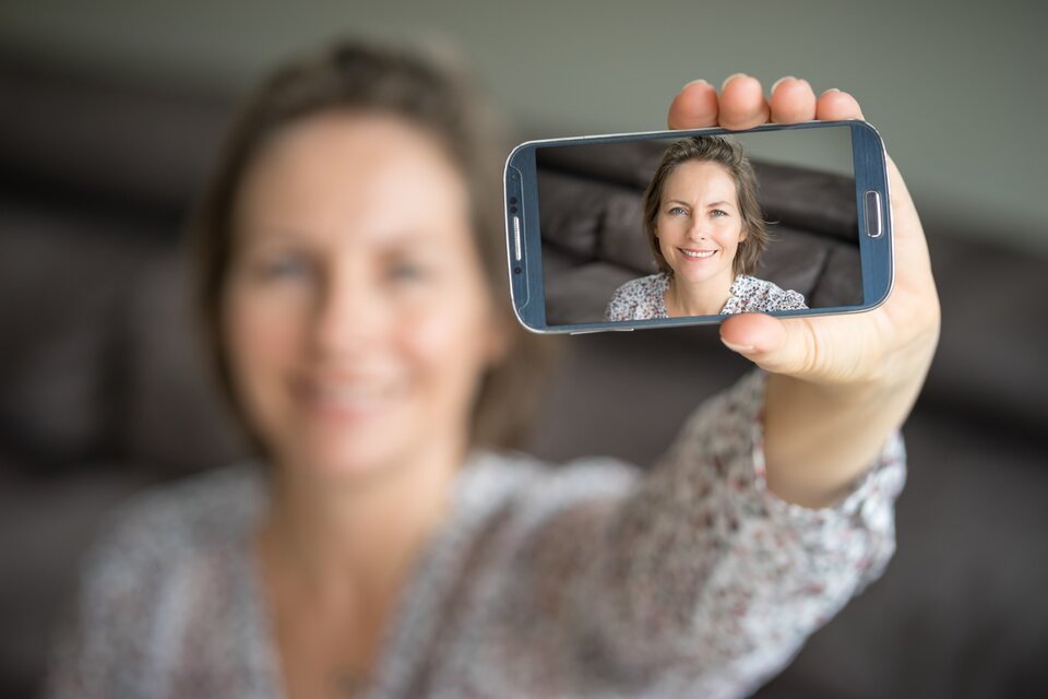 Zdjęcie przedstawia kobietę, która trzyma smartfon przed sobą, robiąc sobie selfie. Ekran telefonu wyraźnie pokazuje jej twarz z uśmiechem, natomiast reszta jej postaci i tło są celowo rozmyte. Kobieta ma jasną cerę i średniej długości włosy, a jej strój to wzorzysta bluzka. Tło wydaje się być wnętrzem mieszkania z kanapą lub sofą. Zdjęcie podkreśla różnicę ostrości między ekranem telefonu a otoczeniem, tworząc efekt głębi.