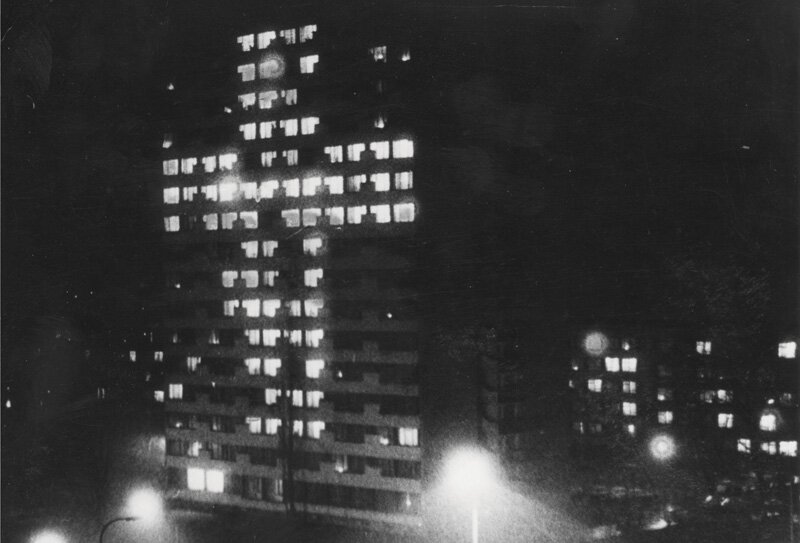 Czarno‑białe zdjęcie nocą przedstawia wysoki budynek mieszkalny, w którym zapalone światła w oknach układają się w kształt krzyża. Przed budynkiem stoją zapalone latarnie. W tle niższe bloki.