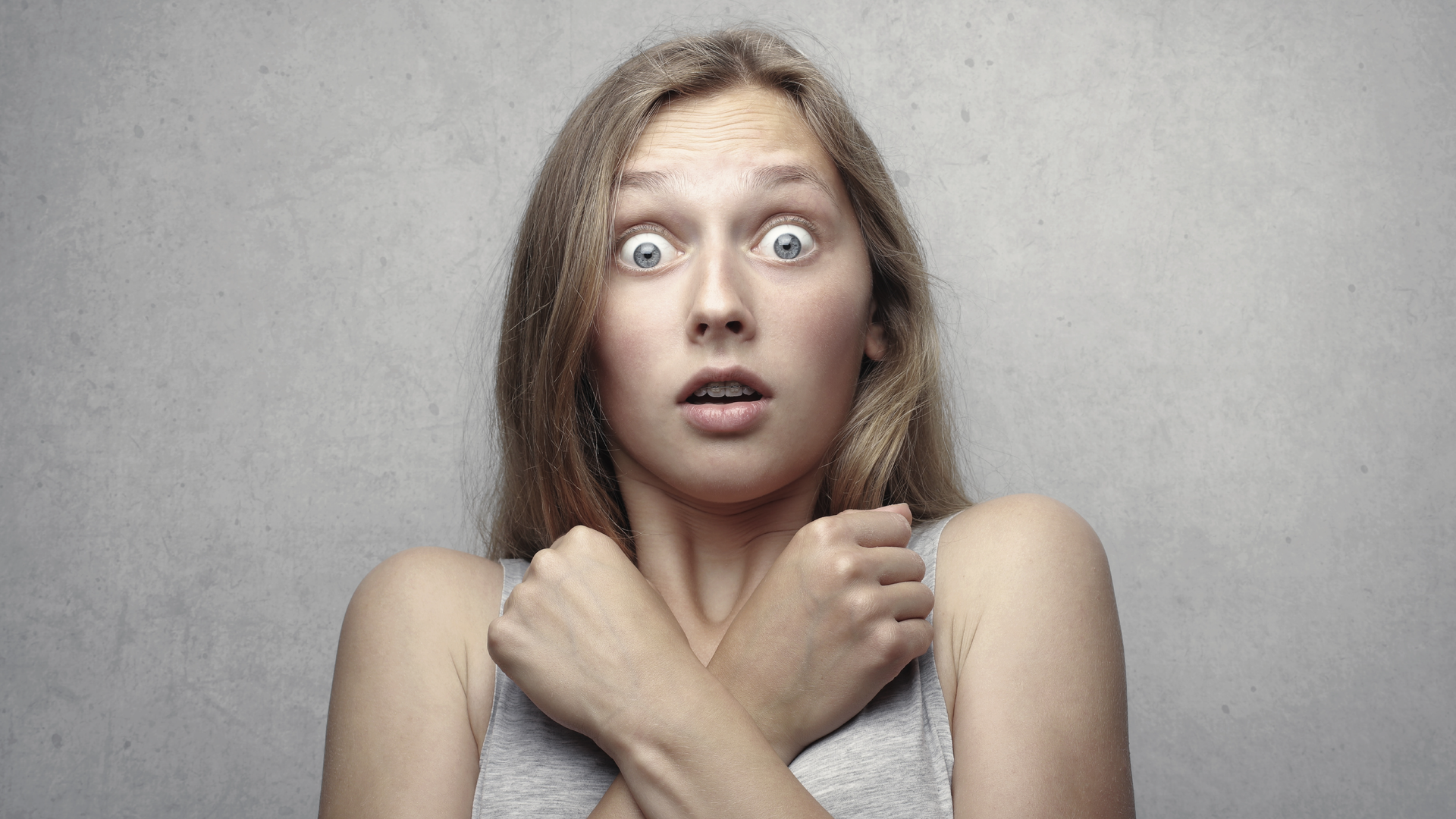 Zdjęcie przedstawia kobietę z szeroko otwartymi oczami i ustami. Jej ręce skrzyżowane są na klatce piersiowej. Zamknięte w pięści dłonie ma skierowane w stronę barków. Kobieta wygląda na przerażoną.