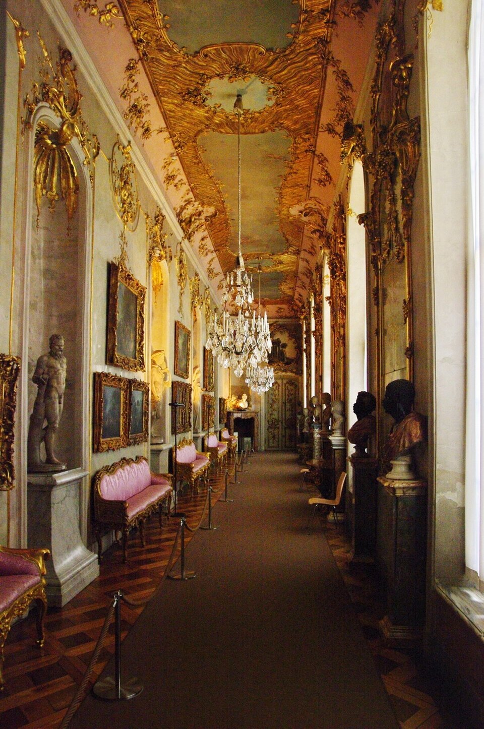 Ilustracja przedstawia wnętrze Pałacyku  Sanssousi w Poczdamie. Ilustracja ukazuje korytarz. Po jednej stronie znajdują się okna, między zbiorami są popiersia na kolumnach. Po prawej stronie, na ścianie wiszą obrazy, między zbiorami są sofy i kolumny. Na kolumnach stoją rzeźby. Sufit pałacu jest bogato zdobiony w ornamenty oraz malowidła.