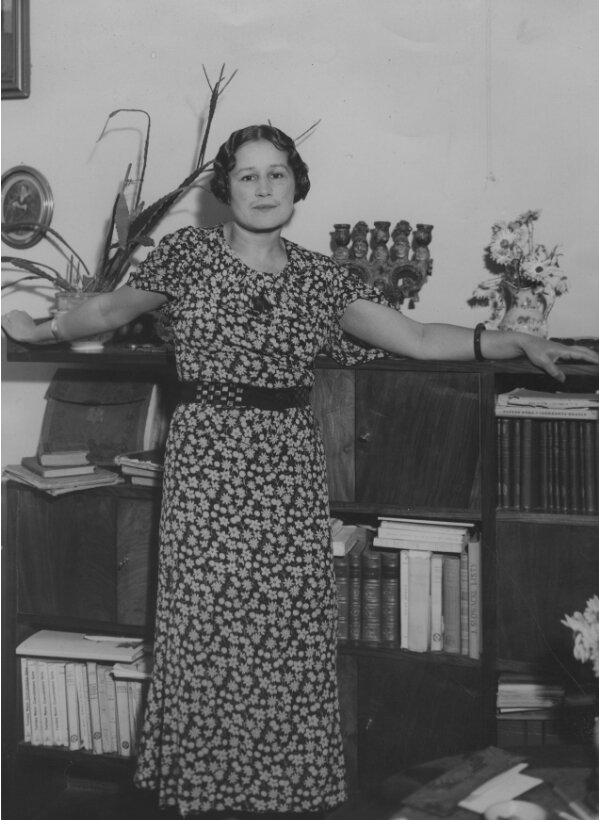 Zdjęcie przedstawia kobietę w średnim wieku. Ma zaczesane, lekko falujące, krótkie włosy. Ubrana jest w długą sukienkę w niewielkie kwiaty. Kobieta wspiera się na regale. Stoi w nim dużo książek. Za plecami kobiety widać kwiaty w wazonie, doniczkę z rośliną, świecznik oraz zdjęcia w ramkach.