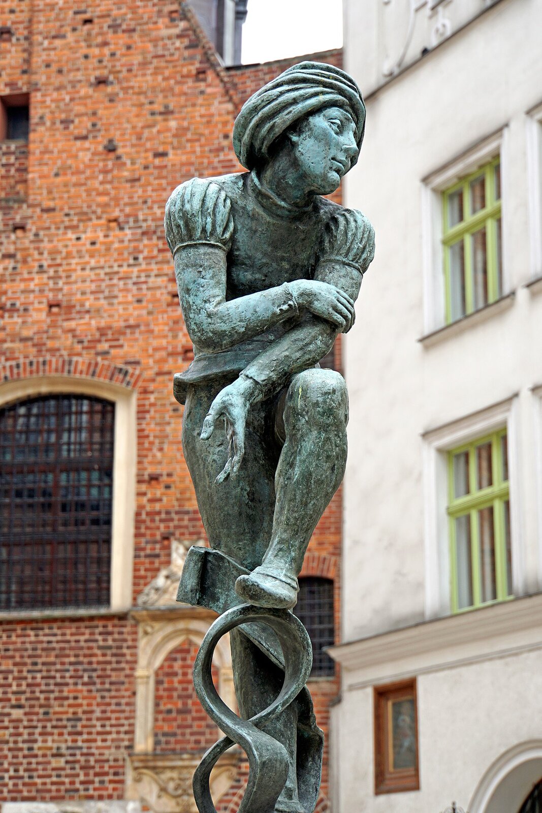 Ilustracja przedstawia figurę młodzieńca, który opiera lewą stopę na podwyższeniu w kształcie cyfry 8. Przedramię lewej ręki spoczywa na zgiętym kolanie. Młodzieniec ubrany jest w obcisłe spodnie, na stopach ma miękkie obuwie, podobne do ciżemek, ale bez wydłużonych nosków. Od góry ubrany jest w koszulę z długimi rękawami, a na wierzch ma nałożony kaftan z krótkimi, bufiastymi rękawkami. Na włosach ma nakrycie głowy z zawiniętego wokół głowy kawałka tkaniny, przypominające turban. W tle ściana domu zbudowanego z czerwonej cegły.