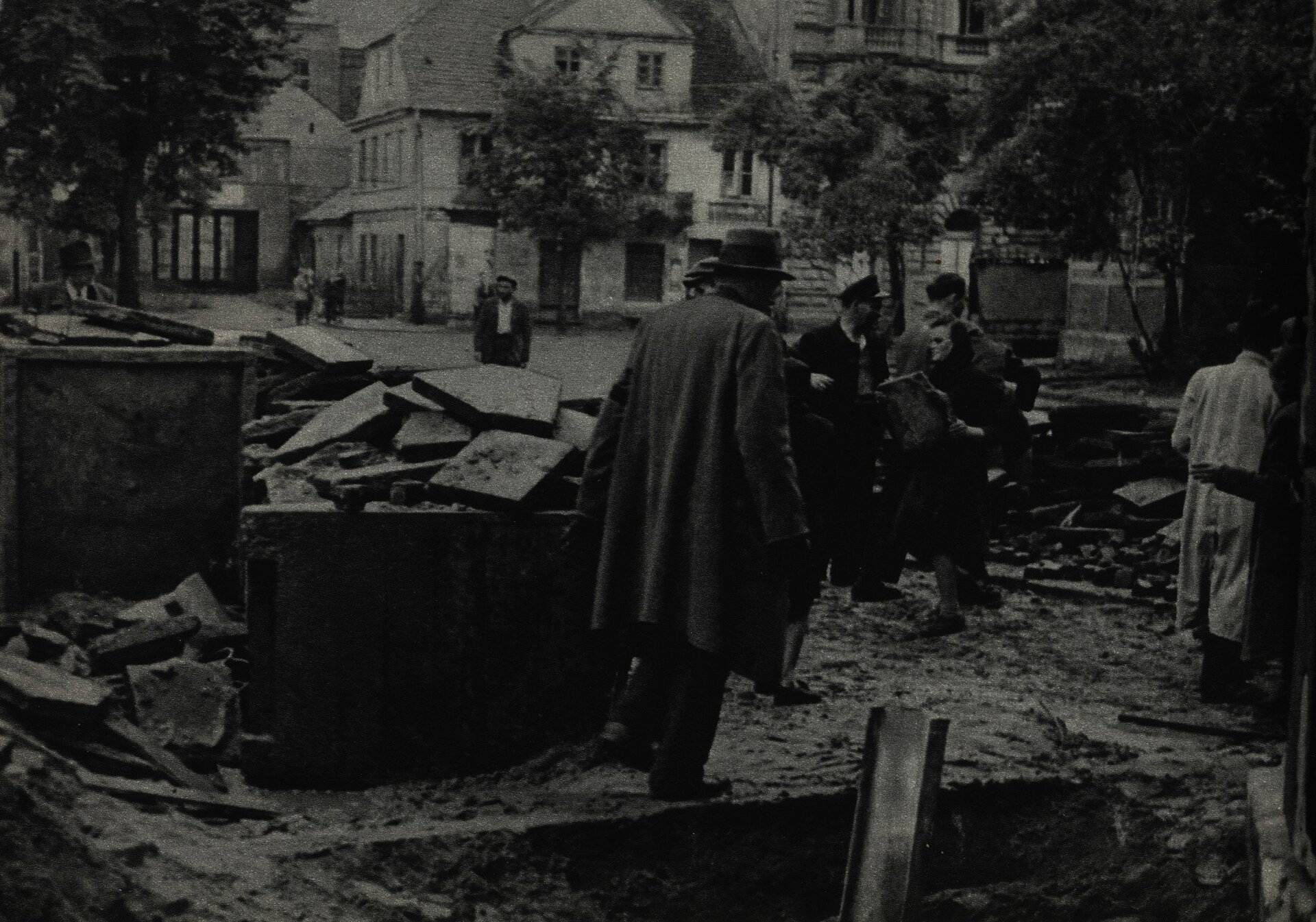 Zdjęcie przedstawia ulicę, na której znajduje się grupa kilku mężczyzn. Biorą oni udział w budowie ulicznej barykady. Jest ona budowana o płyt chodnikowych. W tle widać zabudowania uliczne oraz drzewa pokryte liśćmi.