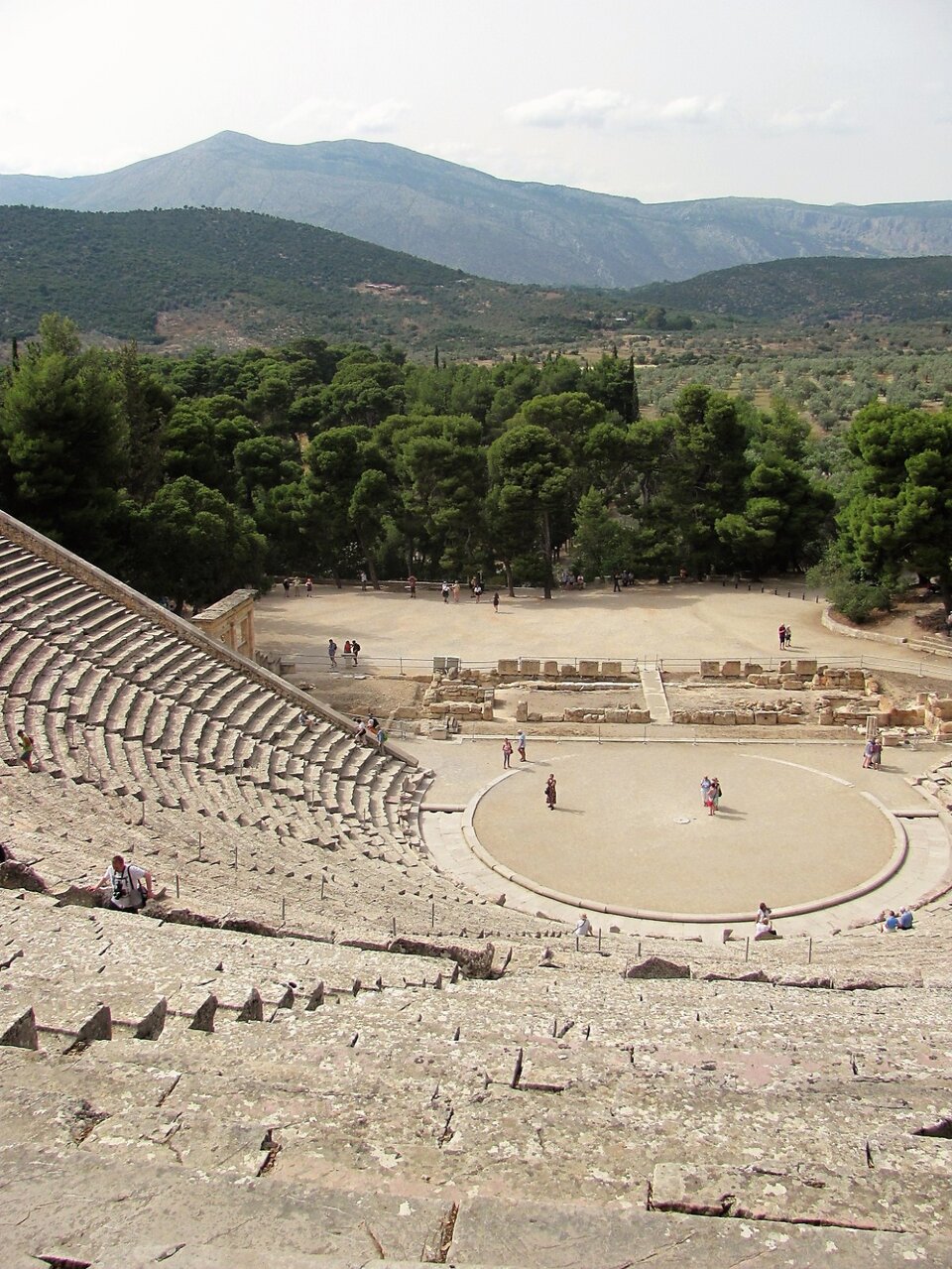 Fotografia przedstawia pozostałości starożytnego teatru w Epidauros. Zdjęcie zostało wykonane z najwyższego miejsca półkolistej schodkowej widowni. Na środku placu znajduje się okrągła orchestra – miejsce dla chóru, za nią szczątki budynków scenicznych. Za teatrem piaszczysty plac i wysokie zielone drzewa. W tyle wysokie wzgórza.