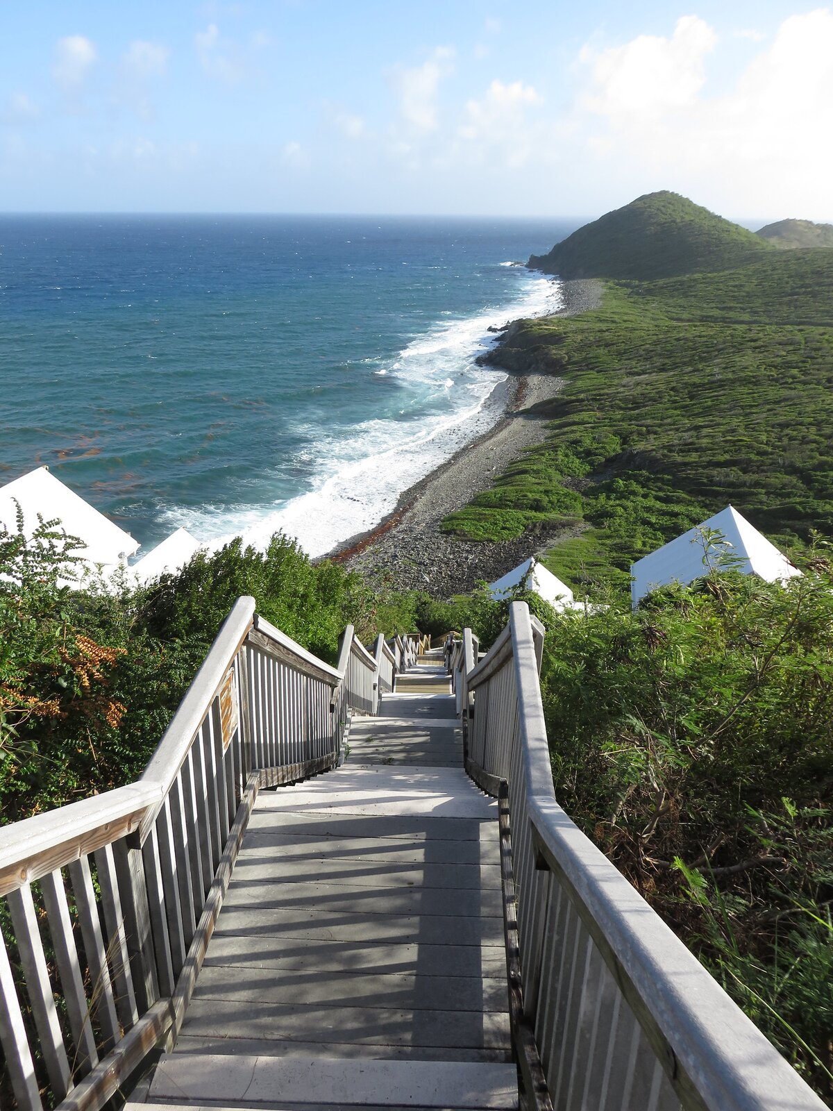 Zdjęcie przedstawia szare, drewniane schody z poręczami po obu ich stronach prowadzące na kamienistą plażę. Dookoła rosną wysokie, zielone krzewy o owalnych listkach. Na dole fotografii znajdują się białe, trójkątne dachy budynków. Poniżej budynków rozpościera się kamienista plaza oraz turkusowe wody morza z białymi falami przy brzegu. Po przeciwnej stronie od morza, znajdują się zielone tereny porośnięte trawą. 