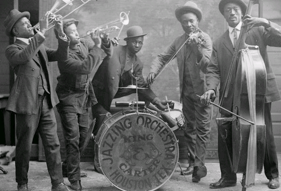 Fotografia przedstawia The King Carter Jazzing Orchestra, Houston 1921 rok. Jest to archiwalne zdjęcie, na którym znajduje się pięciu, zadowolonych, ciemnoskórych mężczyzn. Mężczyźni ubrani są w garnitury, na głowie mają kapelusze. Każdy z mężczyzn grał na innym instrumencie, były to: puzon, trąbka, perkusja, skrzypce i bas.