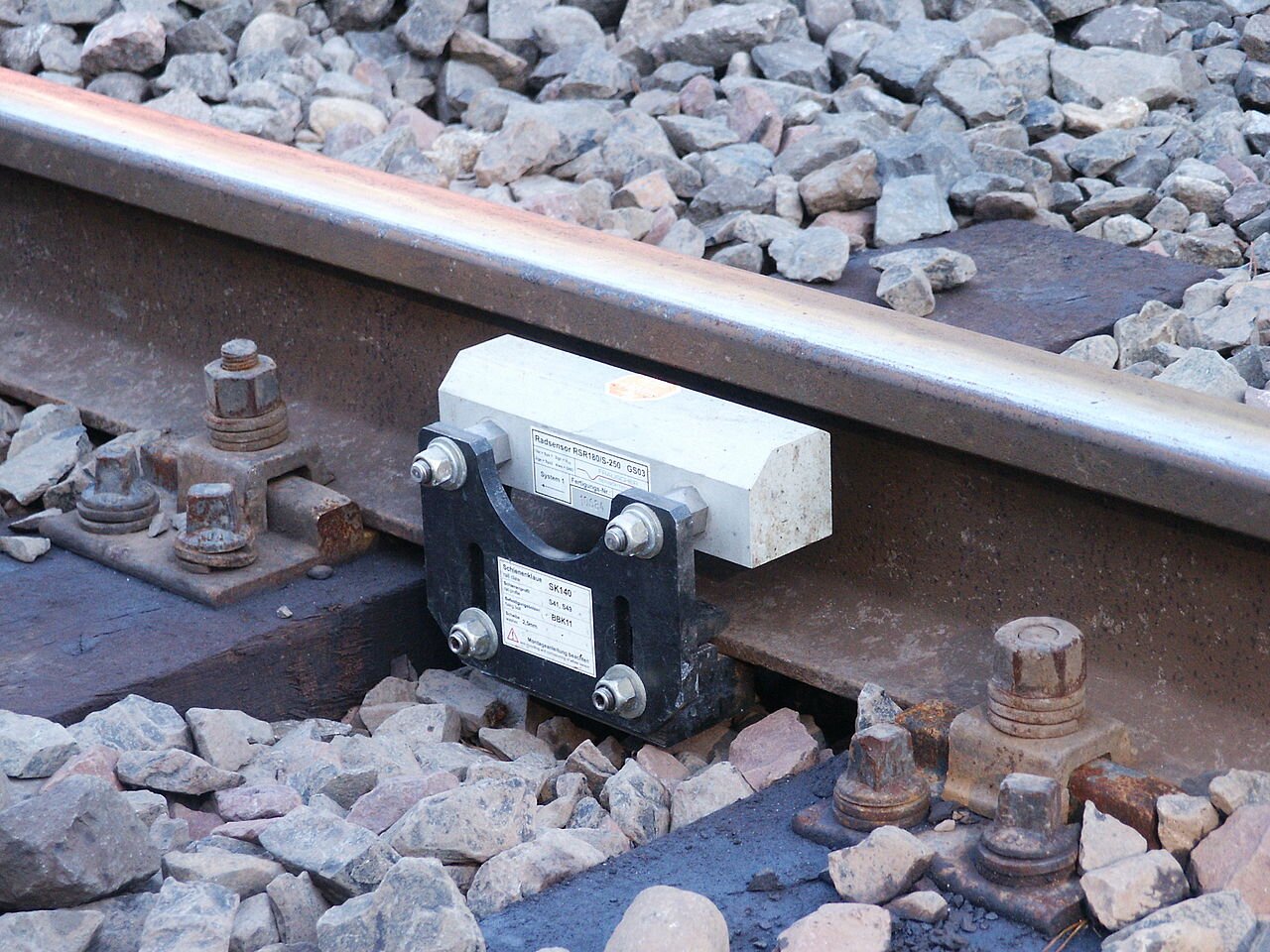 Zdjęcie przedstawia srebrny licznik osi zamontowany do torowiska. Składa się z prostokątnej głowicy i dolnego uchwytu, który wsuwa się w tory. Głowica z jednej strony przykręcona jest do blaszki, za pomocą której połączona jest z dolnym uchwytem, z drugiej na dwóch prętach znajduje się mniejsza część, która po zamontowaniu będzie przylegała do wewnętrznej części torów.