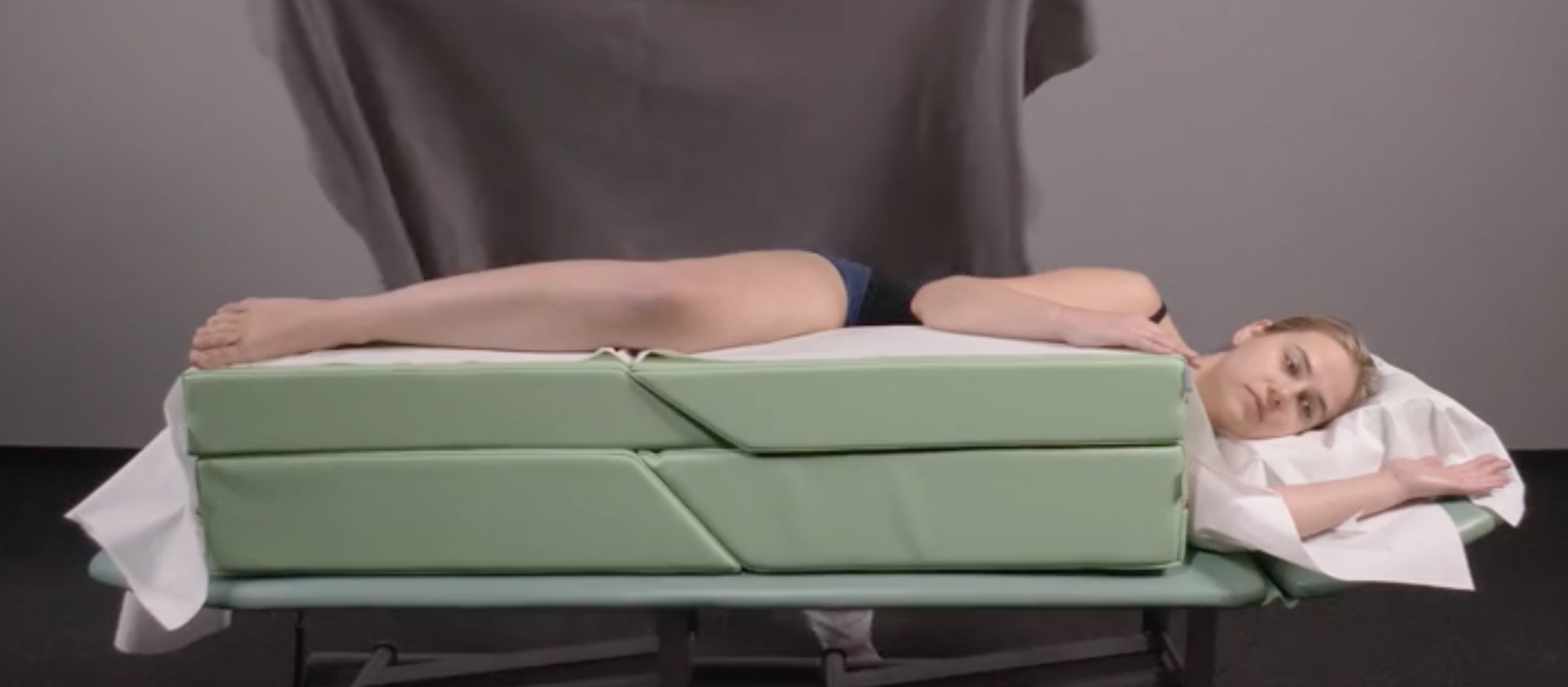 Zdjęcie przedstawiające ułożenie pacjenta w pozycji leżenia bokiem. Pacjent leży na lewym boku, głowę ma podpartą zagłówkiem i poduszką, która przykryta jest prześcieradłem. Lewa ręka leży zgięta na stole do masażu. Prawa ręka i prawa noga pacjenta leżą zgięte na górnej powierzchni klinów ze ścięciem, ułożonych wzdłuż krawędzi stołu do masażu.