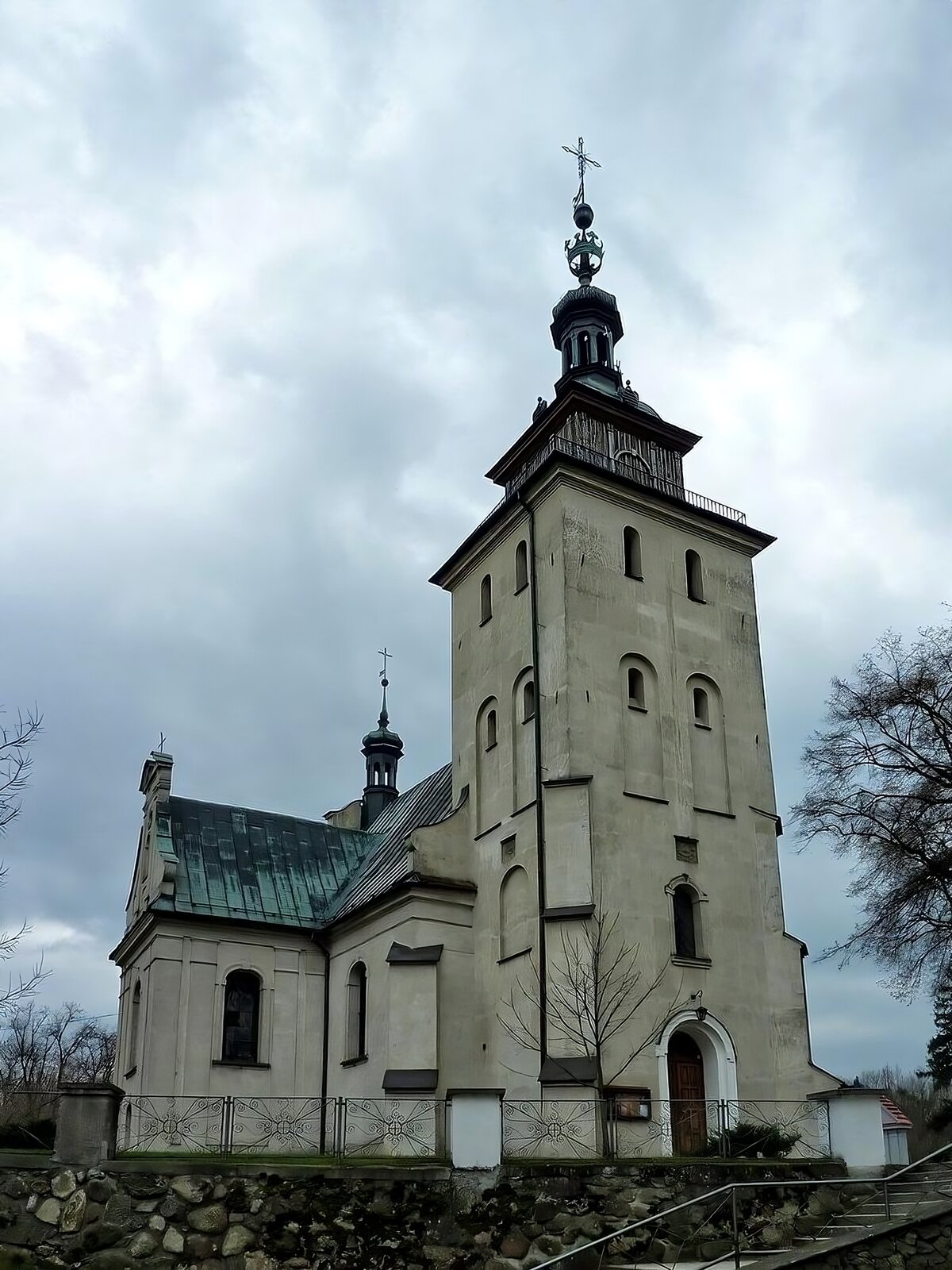 Zdjęcie przedstawia przód kościoła. Na pierwszym planie widoczna jest wysoka wieża, w której znajdują się symetrycznie ułożone okna. Na czubku wieży znajduje się mały balkon ogrodzony czarną, metalową barierką. Czubek jest drewniany, na którym spoczywa metalowa, zielona kopuła. Do kościoła prowadzą drewniane, półokrągłe drzwi. Z prawej strony kościoła widoczne są dodatkowe budowle kościoła - nawa boczna, której dach jest zielony i skośny. Teren kościoła jest ogrodzony metalowym płotem. Do kościoła prowadzą schody widoczne u dołu zdjęcia. Niebo jest zachmurzone. Zdjęcie zostało wykonane w świetle dziennym.