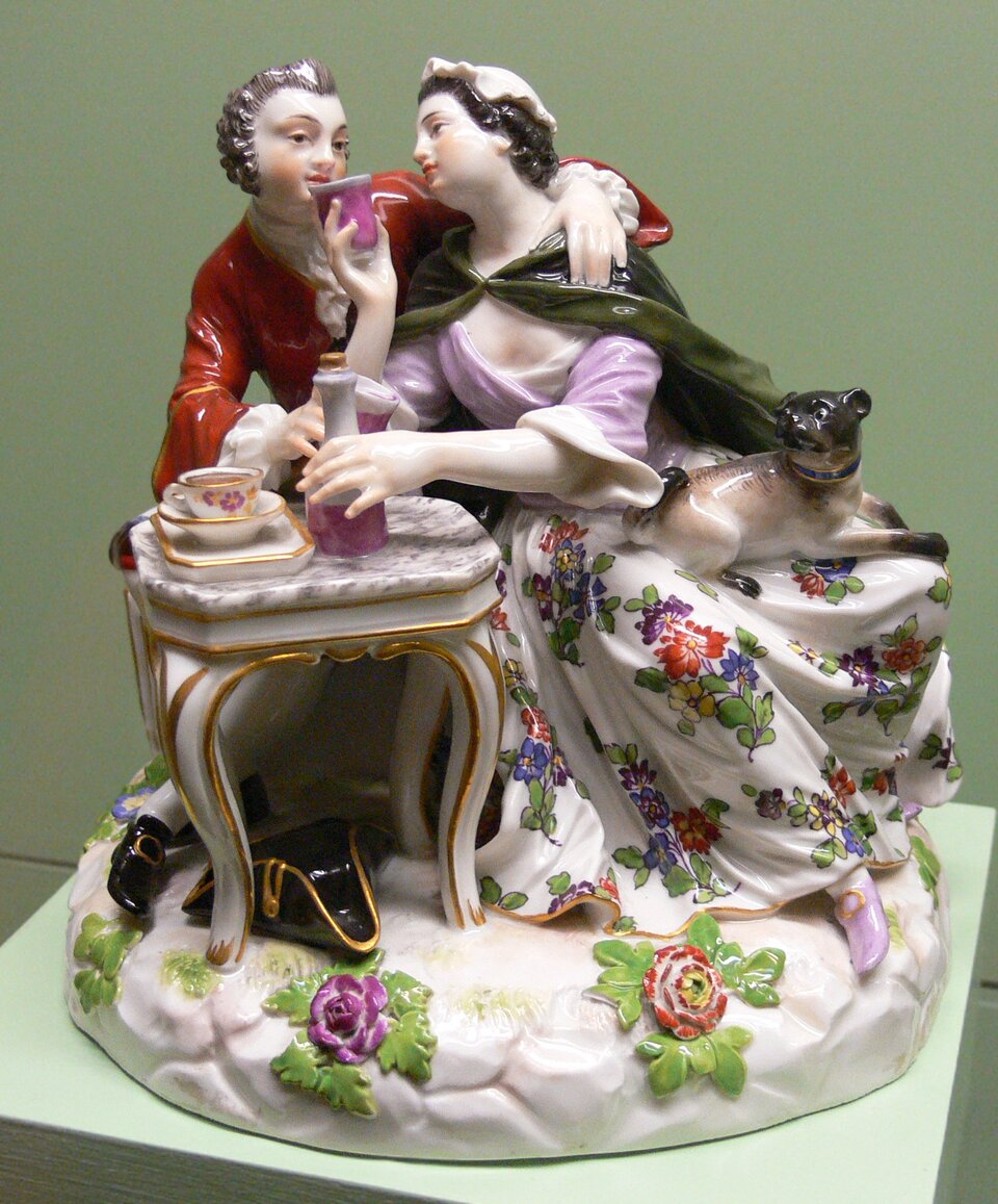 Ilustracja przedstawia figurkę porcelanową zaprojektowaną przez Johanna Joachima Kändlera. Jest to para zakochanych przy stole śniadaniowym. Postacie są ubrane w barwne stroje z epoki. Figurka została wykonana z porcelany miśnieńskiej, jest szkliwiona na biało, a wszystkie szczegóły postaci, ich ubrania oraz detale dekoracyjne są malowane w wielu kolorach. Występują też delikatne złocenia na krawędziach małych elementów.