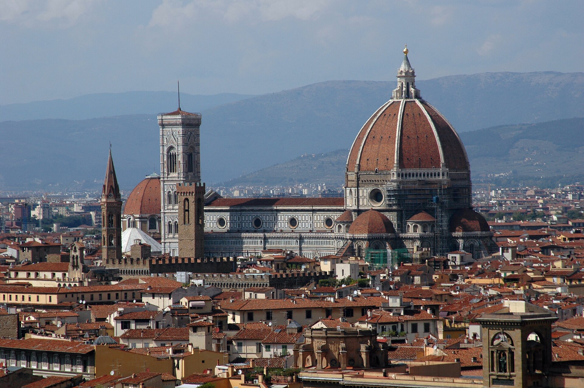 Panorama Florencji. Na zdjęciu bardzo gęsta zabudowa miejska. Czerwone dachy, pośrodku duża katedra Santa Maria del Fiore z czerwonymi kopułami, kilkoma wieżami i bogato zdobioną elewacją. W tle pasmo górskie.