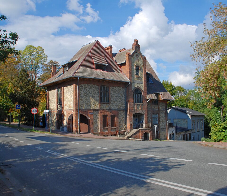Zdjęcie przedstawia dom stojący przy ulicy, którego ściany zewnętrzne wykonane są z równo ułożonego kamienia. Dom ma spadziste dachy. Wybudowany jest na pochyłej działce.  