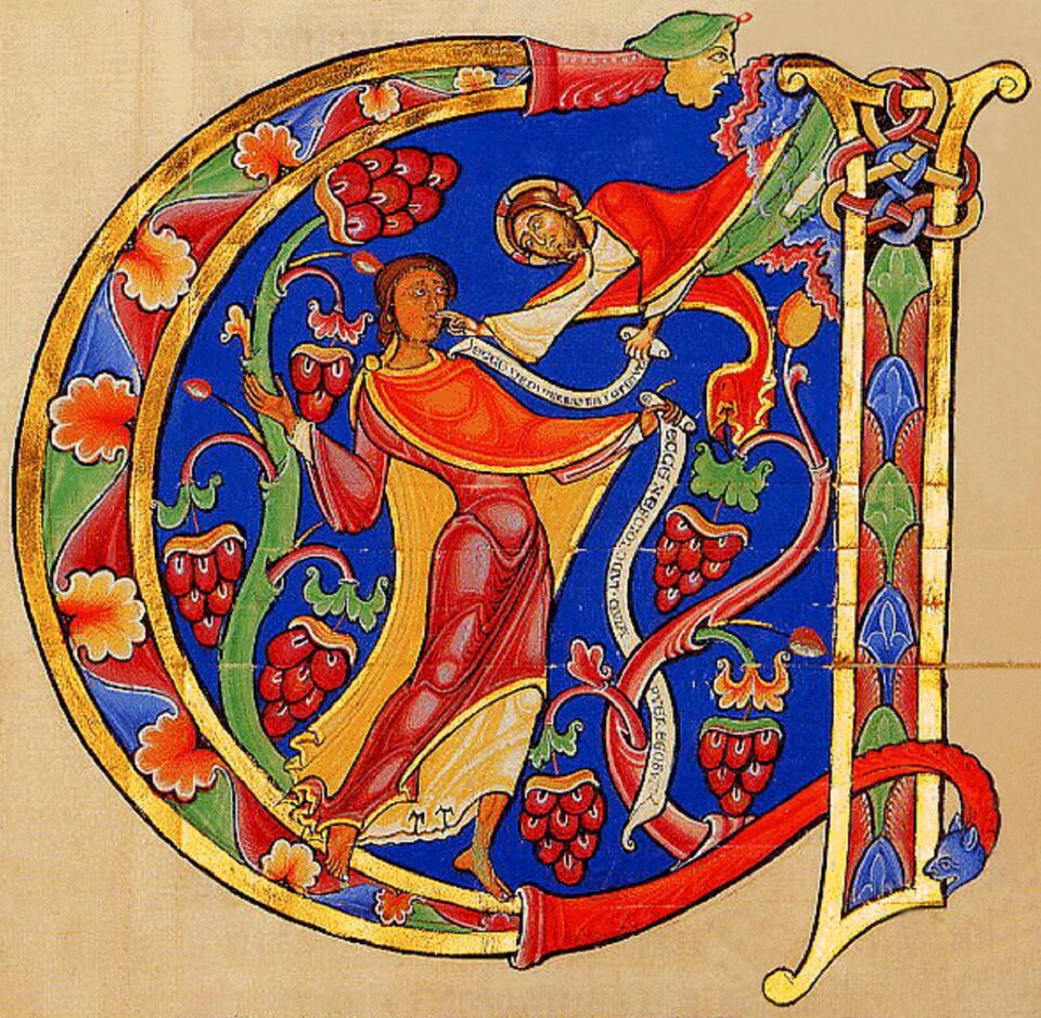 Bogato iluminowany inicjał w kształcie litery, wypełniony sceną figuralną na intensywnie niebieskim tle. Centralna postać w długiej, czerwono‑złotej szacie stoi wśród wijących się pędów winorośli z kiściami owoców, trzymając gałązkę. Z prawej strony ku postaci pochyla się druga figura, wyłaniająca się z ornamentu, z wstęgą zawierającą napis. Całość otaczają dekoracyjne motywy roślinne i geometryczne w żywych barwach czerwieni, zieleni, błękitu i złota, charakterystyczne dla iluminacji średniowiecznych rękopisów.