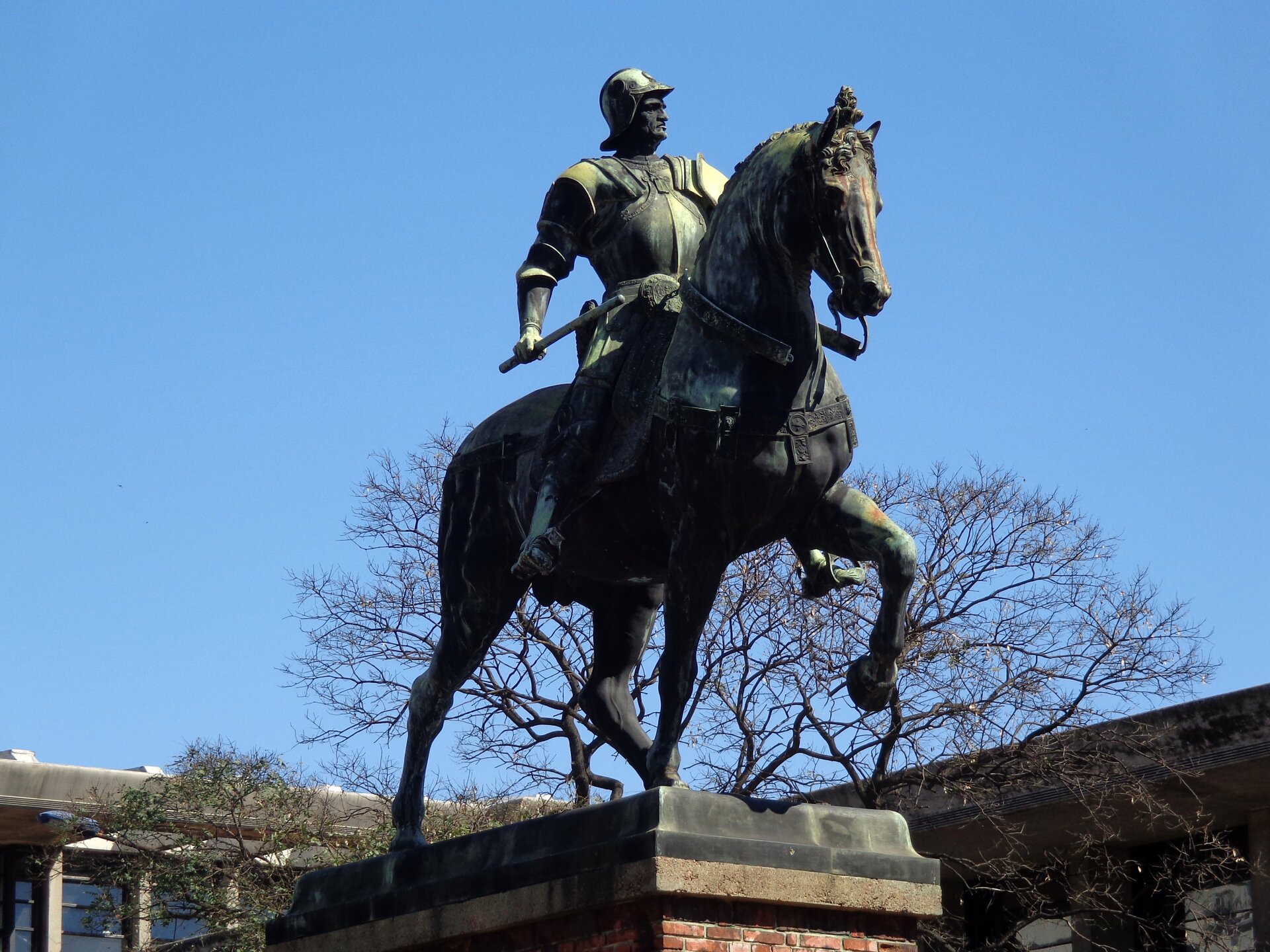 Zdjęcie przedstawia rzeźbę w zielonkawych odcieniach na tle niebieskiego nieba. Posąg stanowi mężczyznę odzianego w zbroję oraz hełm, który jedzie galopem na nieustraszonym koniu. W swojej prawej ręce trzyma długi przedmiot.
