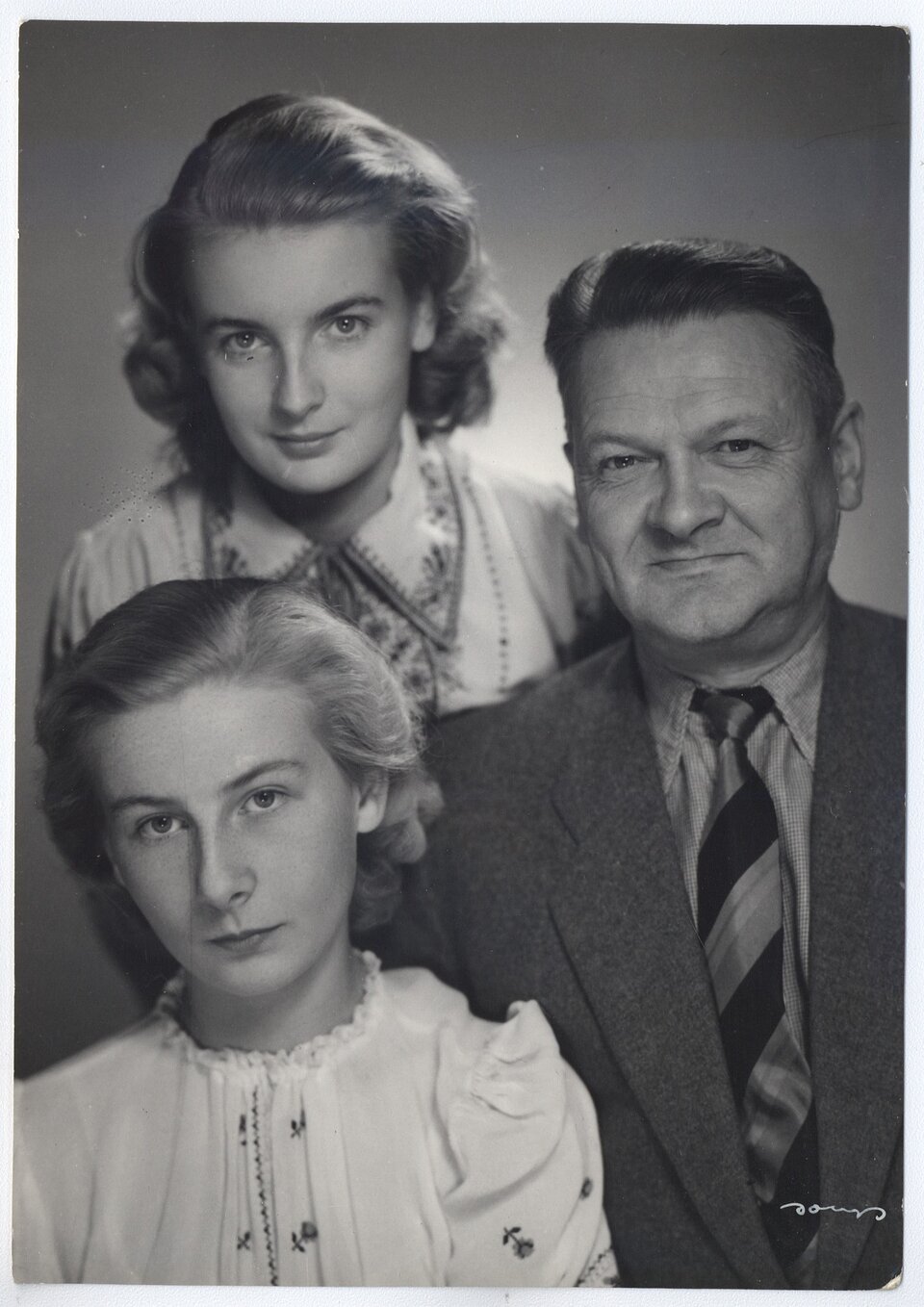 Czarno‑białe zdjęcie portretowe trojga ludzi. Po lewej dwie młode kobiety o jasnych włosach sięgających ramion. Kobieta znajdująca się poniżej ma na sobie białą bluzkę ozdobioną haftowanymi kwiatkami, z bufiastymi rękawkami, wiązaną pod szyją na tasiemkę. Pochyla lekko głowę w lewą stronę, ma poważny wyraz twarzy. Kobieta znajdująca się powyżej ma na sobie zdobioną kwiatowym haftem bluzkę z kołnierzykiem. Lekko uśmiecha się. Po prawej znajduje się uśmiechnięty mężczyzna w średnim wieku. Jego włosy są ciemne, krótkie i zaczesane do góry. Ma na sobie koszulę z kołnierzykiem, krawat w ciemne, skośne pasy i ciemną marynarkę.