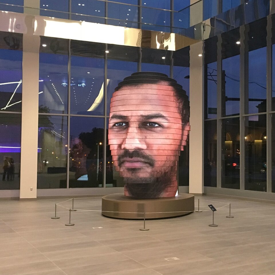 Zdjęcie przedstawia interaktywną rzeźbę głowy. Rzeźba składa się z podłużnych paneli LED, ułożonych w poprzek, jeden na drugim. Na tych panelach wyświetlany jest obraz, który tworzy ludzką twarz. Przedstawiony został mężczyzna w średnim wieku. Ma duży nos, lekko rozchylone usta, ciemny zarost. Oczy ciemne, głęboko osadzone. Na głowie ma krótkie, ciemne włosy. Rzeźba oddaje nawet światło, które odbija się od twarzy, dzięki czemu jest bardzo realistyczna. Stoi na okrągłej podstawce, otoczona barierkami, w dużym, oszklonym pomieszczeniu.