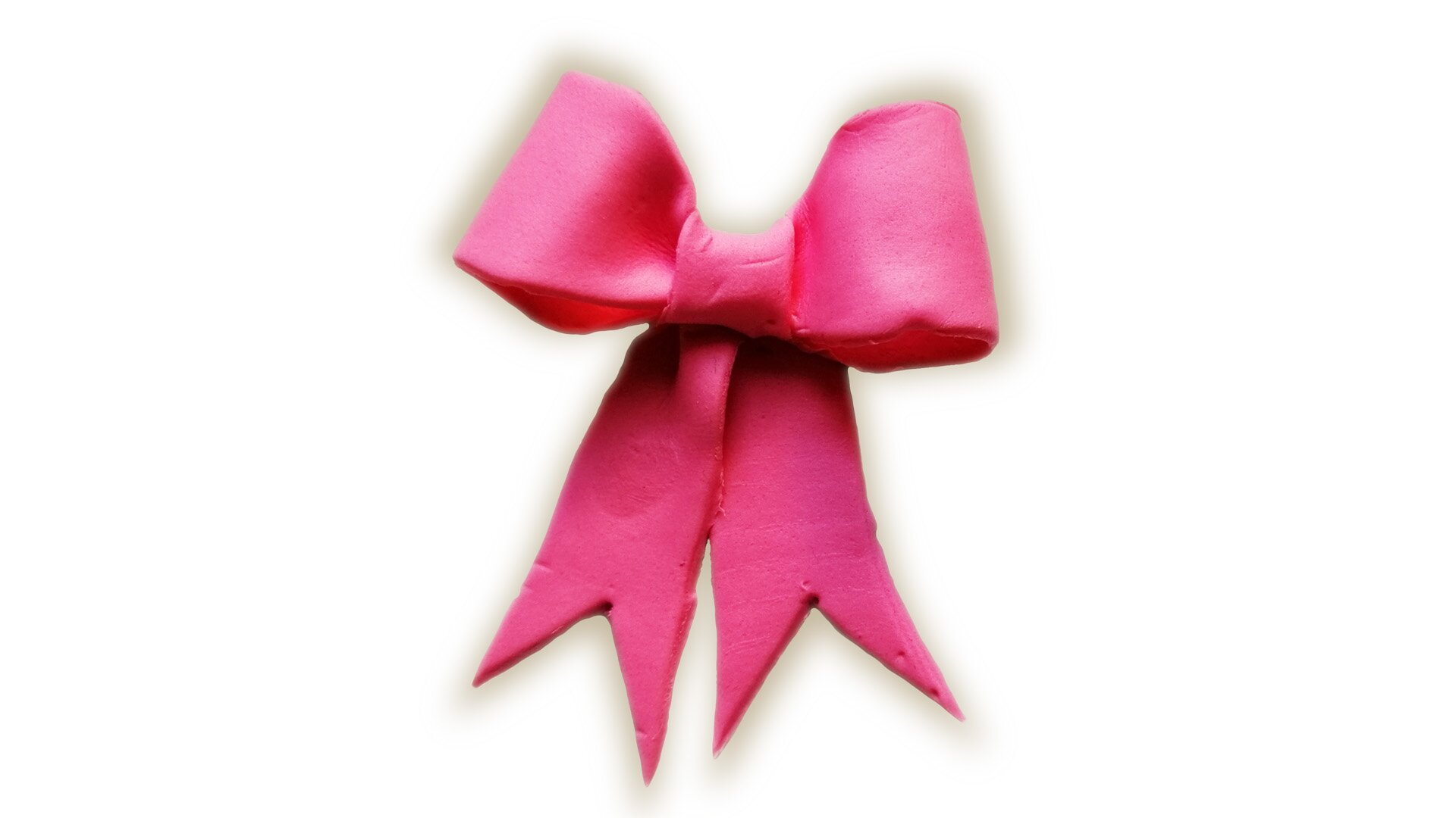 Zdjęcie przedstawia kokardę z masy cukrowej. Widoczna jest różowa kokarda z zakończeniami opadającymi w dół, z wyciętymi ząbkami. 