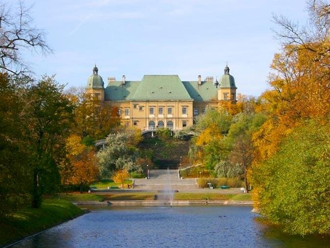 Na zdjęciu zbiornik wodny z fontanną. W głębi zdjęcia, na wzgórzu, znajduje się dwupiętrowy zamek, po bokach którego są dwie wieże. Za zamkiem oraz po bokach akwenu zielono‑pomarańczowe drzewa. Za akwenem schody na wzgórze. 