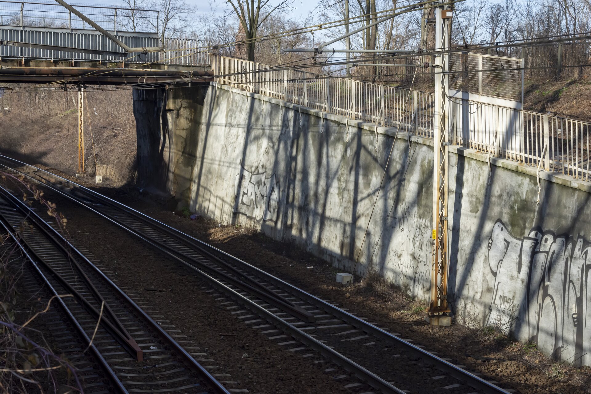 Zdjęcie przedstawia mur oporowy. Jest on wybudowany z betonu i ciągnie się wzdłuż torów kolejowych, zapewniając stabilność i bezpieczeństwo dla ruchu kolejowego. Obok muru oporowego znajduje się most dla samochodów. Na moście, jak i na murze oporowym znajduje się barierka ochronna, zapewniająca bezpieczeństwo dla użytkowników. 