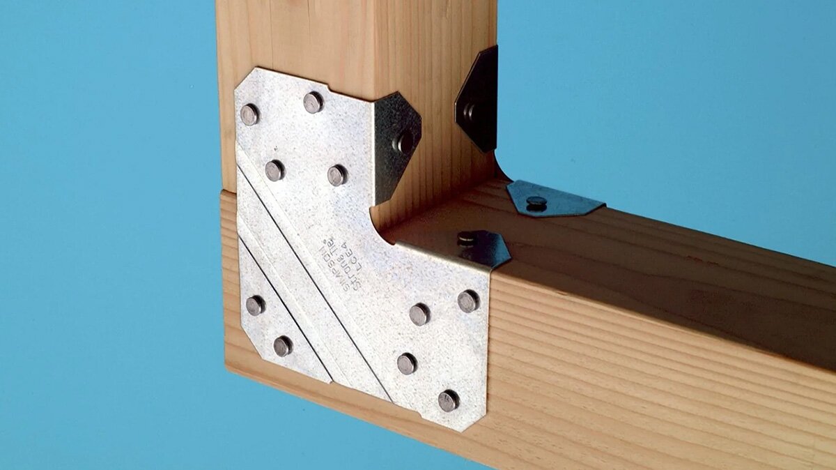 Na zdjęciu widoczny drewniany element tworzący kąt prosty, przypominający róg mebla lub ramy, na nim znajduje się przymocowana płasko metalowa blaszka z kilkoma wypustkami. Widać, że z drugiej strony drewnianej części znajduje się taka sama blaszka.