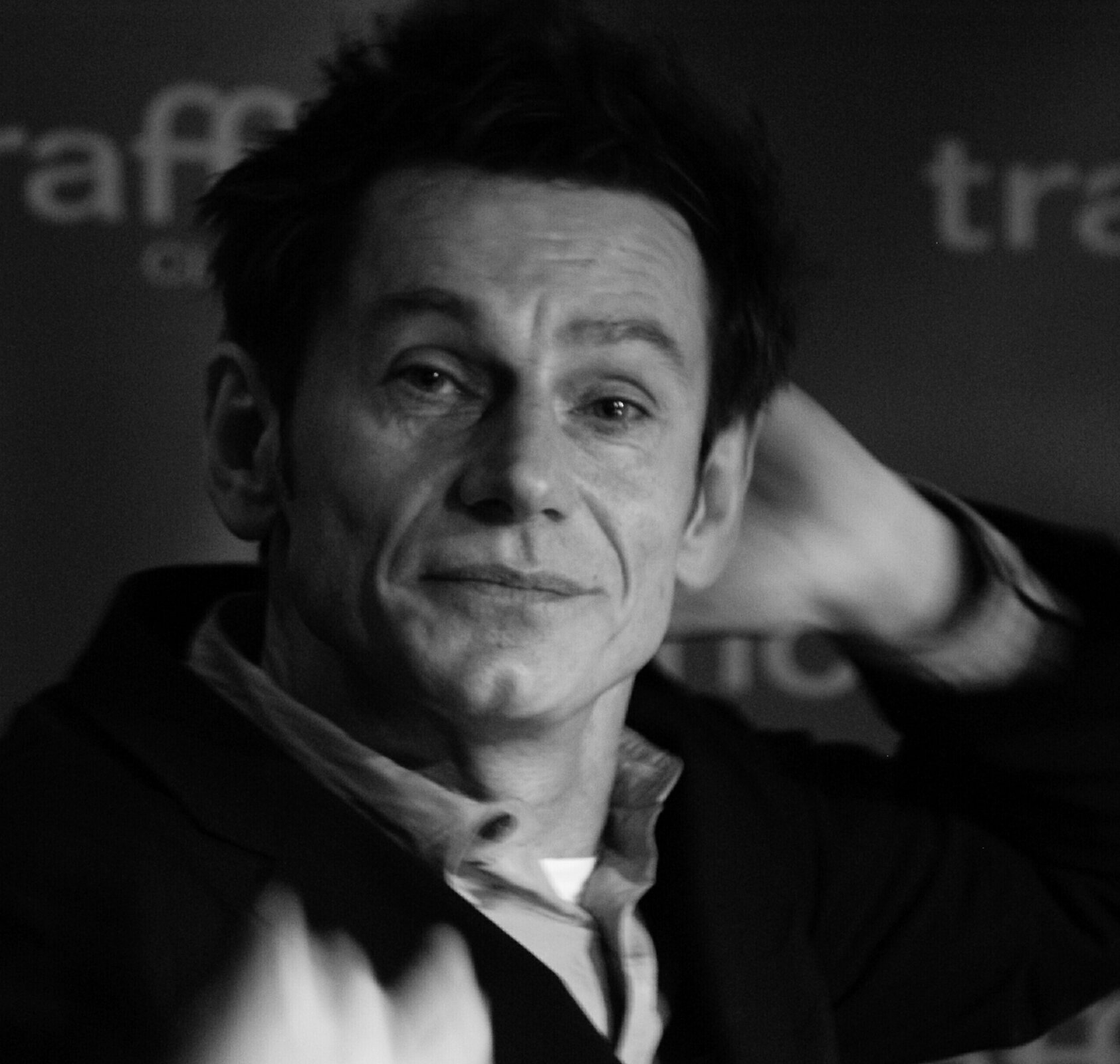 Zdjęcie przedstawia twarz dojrzałego mężczyzny. Widoczne są na niej zmarszczki. Mężczyzna ma kwadratową twarz, wydatny nos, wysokie czoło. Jego włosy są ciemne, proste, zwichrzone. Mężczyzna ubrany jest w podkoszulek, koszulę i marynarkę.