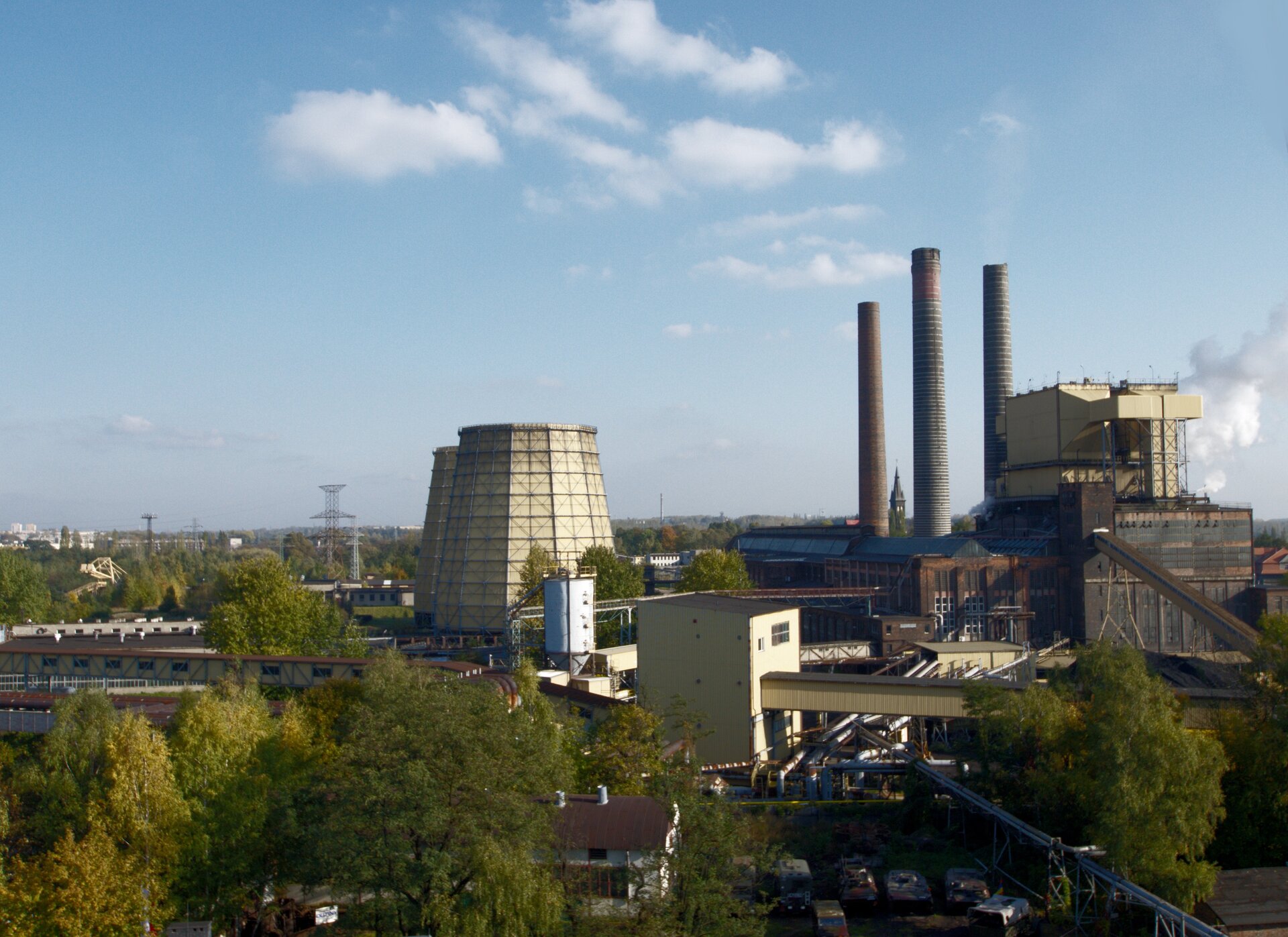 Zdjęcie przedstawia elektrociepłownię w Zabrzu. Na jej terenie znajdują się: trzy kominy, budynki ciepłowni, linie transportujące, dwa silosy oraz kilka drzew.
