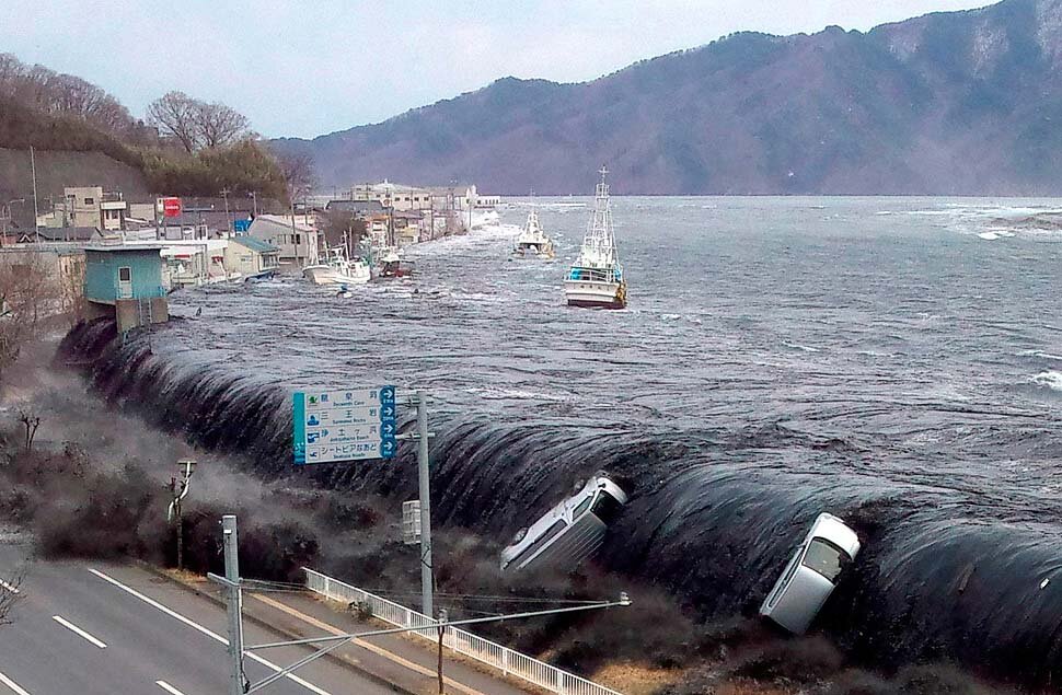 Zdjęcie przedstawia tsunami w Japonii. Z lewej strony znajduje się ulica, na którą przelewają się wody tsunami z prawej strony. Woda porwała ze sobą samochody. Po wodzie pływają niewielkie statki. W tle znajduje się góra.