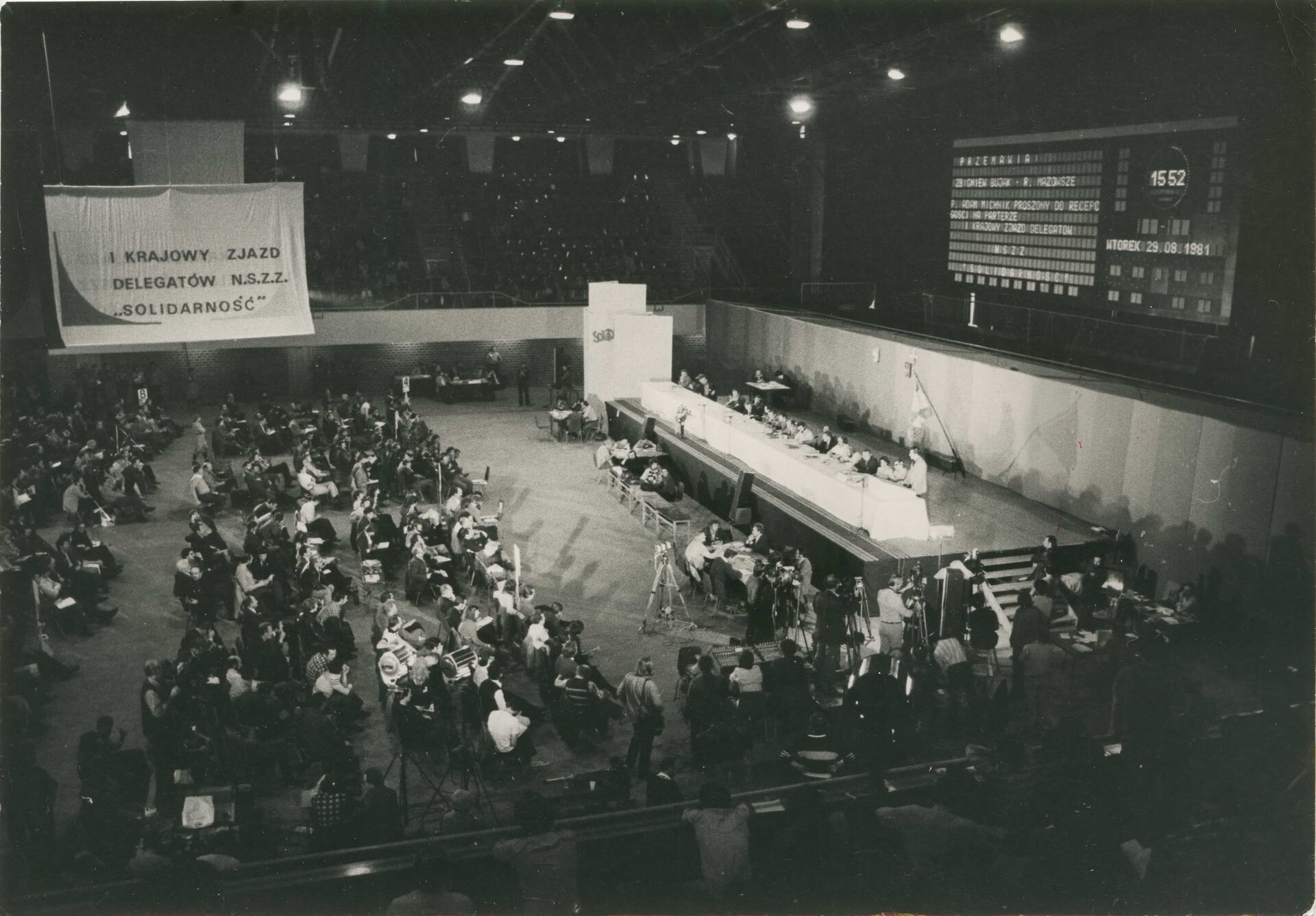 Ilustracja przedstawia I Krajowy Zjazd Delegatów NSZZ „Solidarność” w Gdańsku, wrzesień 1981 roku. Na zjazd zostało wybranych 896 delegatów z 38 regionów. Ludzie siedzą na krzesłach na dużej hali, przed nimi znajduje się podwyższenie i stół, za którym zasiadają przemawiający. 