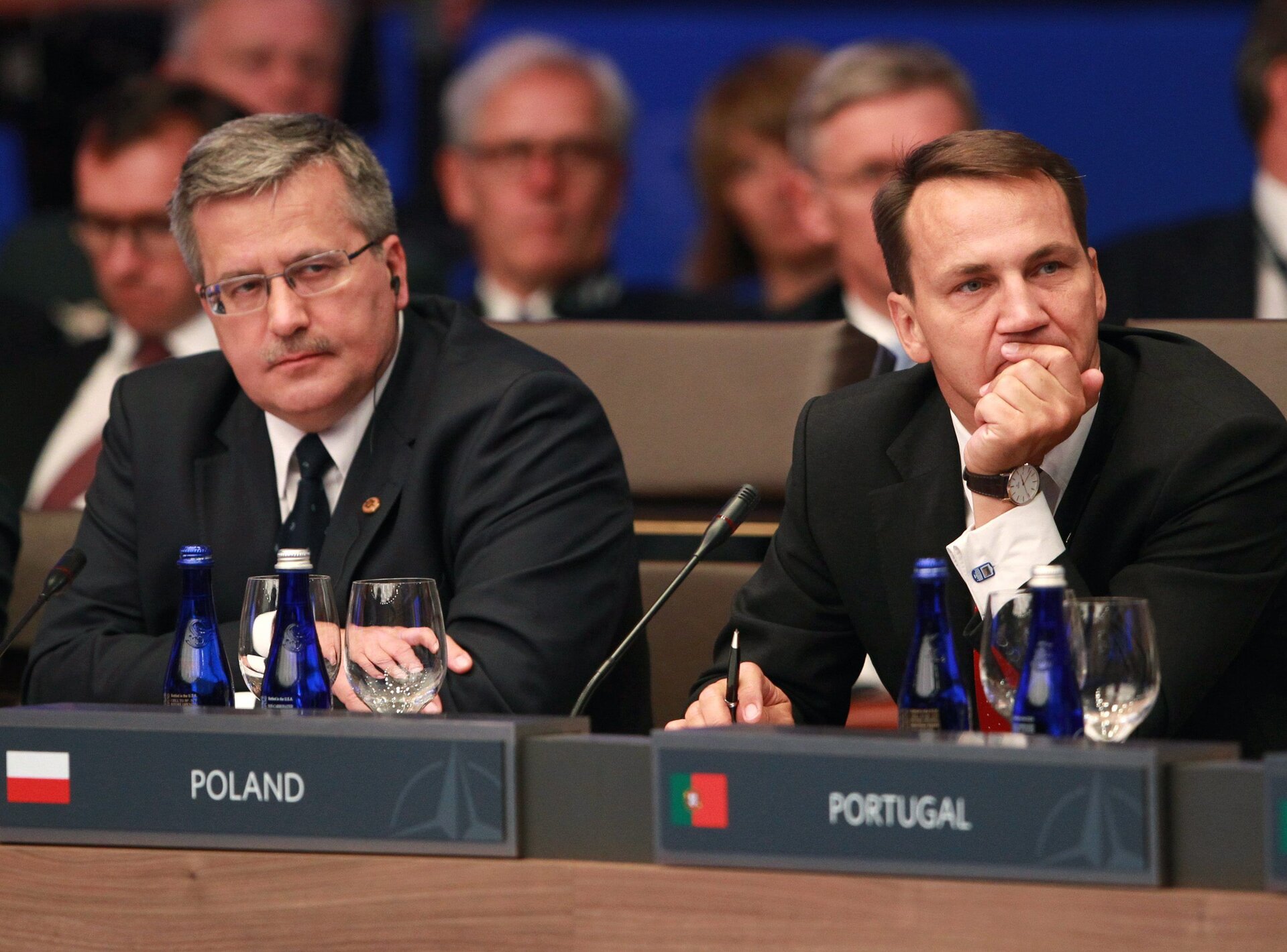 Zdjęcie przedstawia Prezydenta Bronisława Komorowskiego wraz z Ministrem Spraw Zagranicznych Radosławem Sikorskim. Siedzą wspólnie na międzynarodowym posiedzeniu. Przed nimi stoją tabliczki z opisami krajów: POLAND i PORTUGAL. W tle liczni politycy. 
