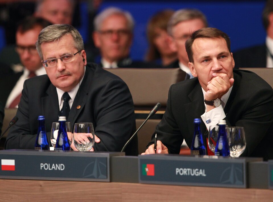 Zdjęcie przedstawia Prezydenta Bronisława Komorowskiego wraz z Ministrem Spraw Zagranicznych Radosławem Sikorskim. Siedzą wspólnie na międzynarodowym posiedzeniu. Przed nimi stoją tabliczki z opisami krajów: POLAND i PORTUGAL. W tle liczni politycy.