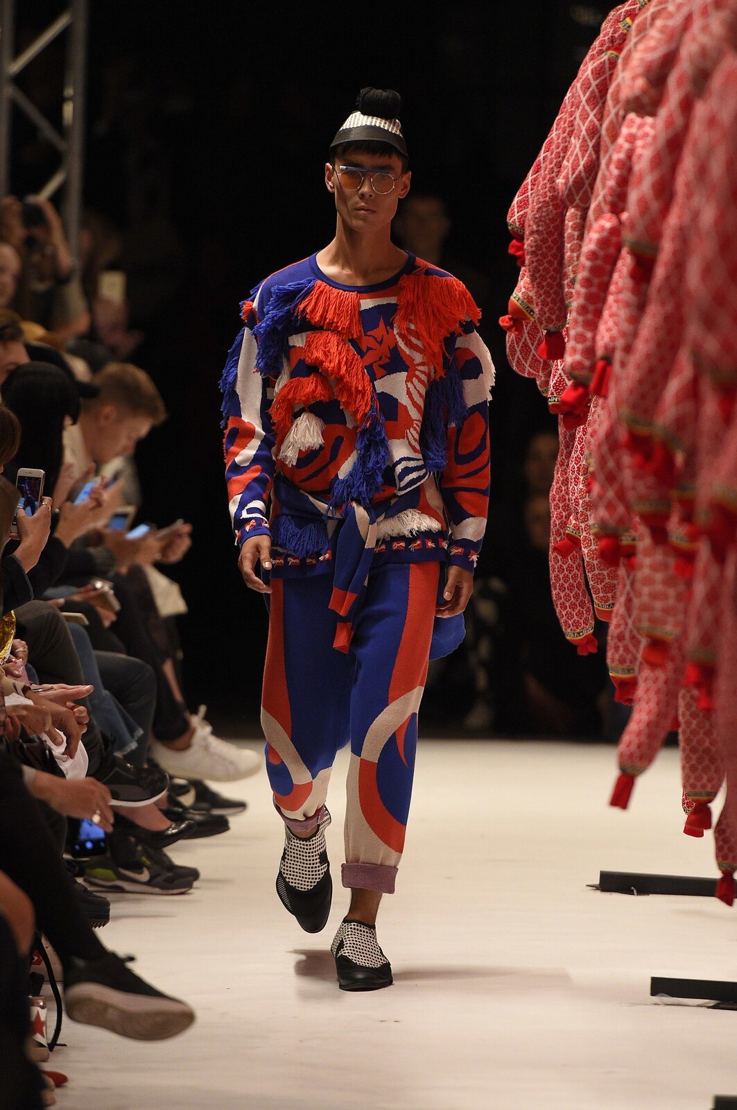 Fotografia przedstawia modela na wybiegu. Mężczyzna ma na sobie spodnie oraz bluzę z materiału wyglądem przypominającego dresową bawełnę w kolorach granatowym, czerwonym oraz białym. Kolory łączą się w nieregularne wzory, widoczne są również przeszycia z wystającymi kawałkami materiału – frędzlami. Buty mężczyzny wydają się nie mieć podeszwy, co sprawia wrażenie, że są bardzo cienkie. Na nosie modela widoczne są okulary w okrągłej oprawie, a na jego głowie czarno‑biała czapka z niewielkim daszkiem oraz pomponem.