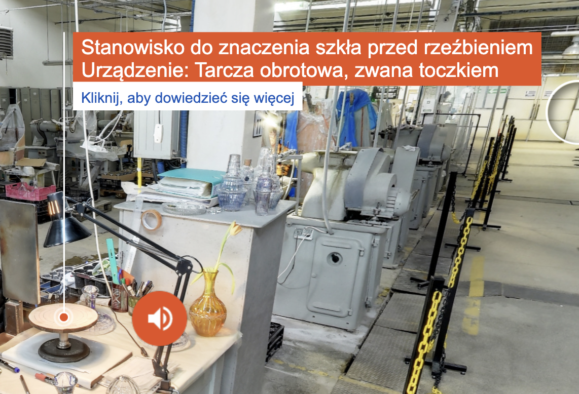 Zdjęcie przedstawia zrzut ekranu z wycieczki wirtualnej. Ukazany jest fragment hali produkcyjnej kryształów. Na zdjęciu zaznaczona jest punktorem tarcza toczka i linia do opisu: Stanowisko do znaczenia szkła przed rzeźbieniem, urządzenie: tarcza obrotowa, zwana toczkiem, kliknij aby dowiedzieć się więcej. W pobliżu punktora znajduje się symbol głośnika.