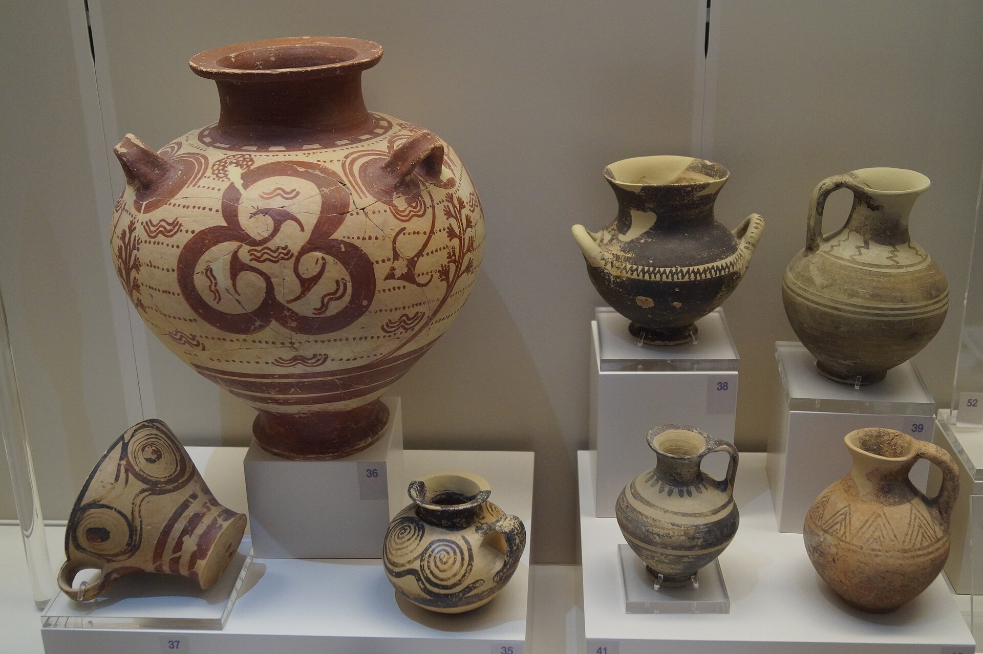 Fotografia autorstwa Schuppi przedstawiająca grupę waz, które zostały odnalezione w mykeńskich nekropoliach. Wazy stoją za szybą, na ekspozycji w muzeum i jest ich siedem (jedyna duża, stojąca z lewej strony) oraz sześć mniejszych (stojące z prawej strony dużej wazy i pod nią); część naczyń stoi na podestach, a część na półkach, jedna z waz leży na boku. Największe naczynie swoim kształtem przypomina bańkę, posiada wąską podstawę oraz szyjkę. Jest pomalowana brązowymi i beżowymi kolorami. W centralnej części dużej wazy znajduje się ornament kształtem przypominającą liść koniczyny. Mniejsze wazy są zdobione wzorami geometrycznymi lub kolistymi.