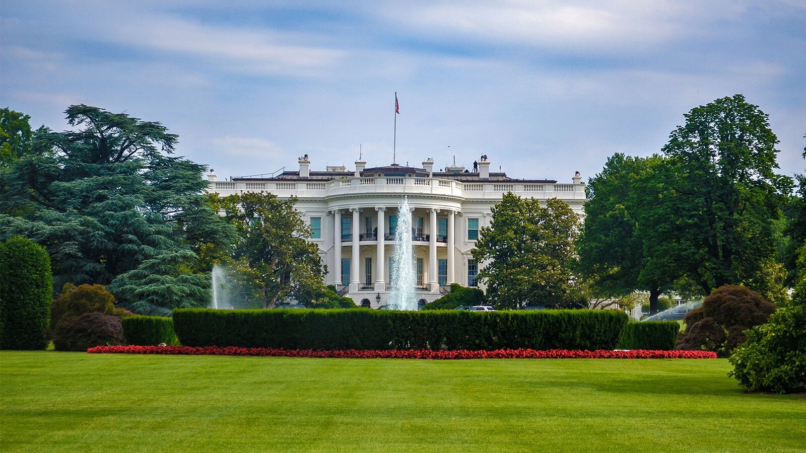 Zdjęcie przedstawia Biały Dom od strony ogrodów. Dwupiętrowy pałac z efektownymi balkonami, na którego dachu widoczna jest flaga amerykańska. Budynek ma białą elewację. Przed budynkiem widoczna fontanna.