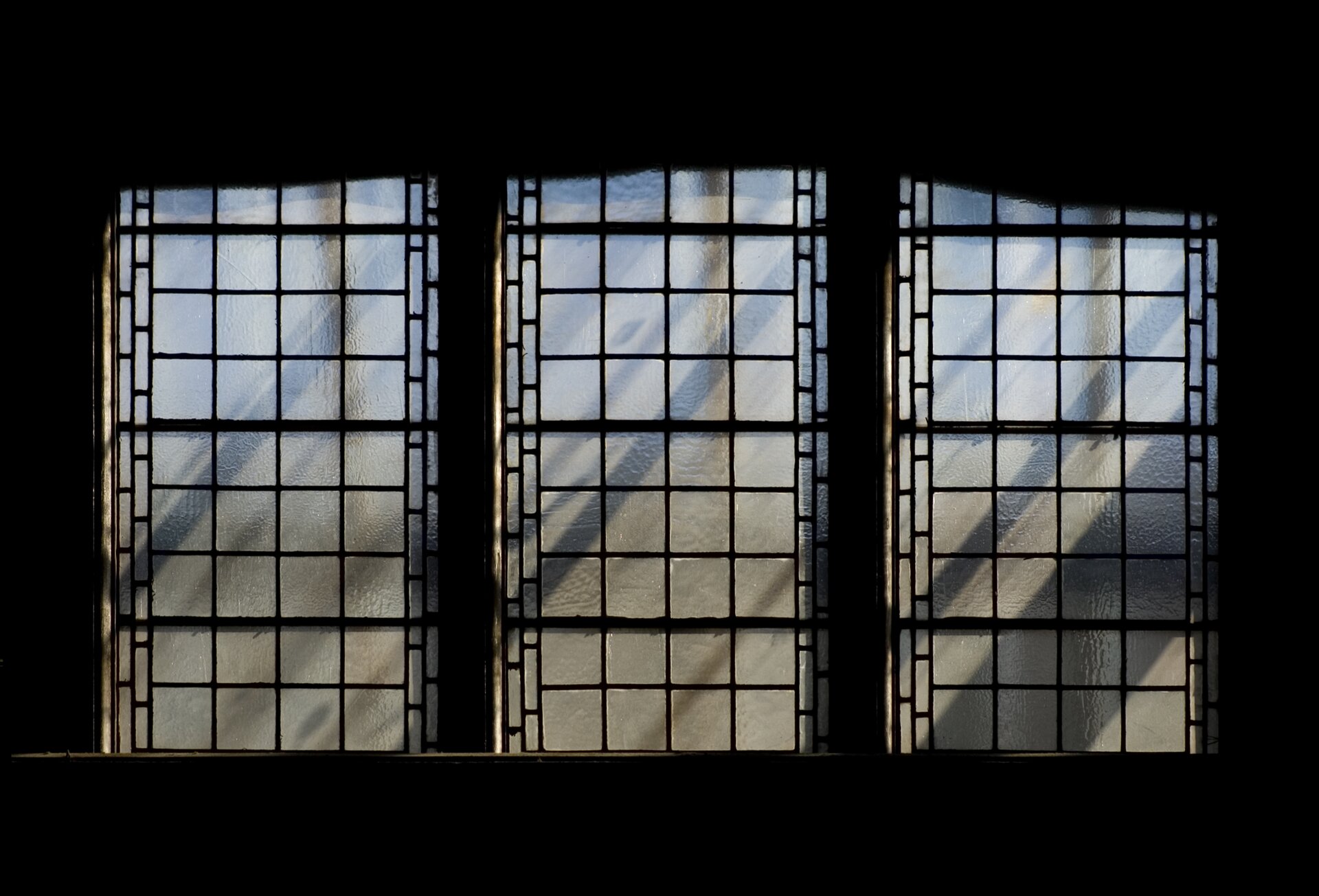 Ilustracja przedstawia refleks świetlny. Ukazuje trzy znajdujące się obok siebie okna, dzielone na mniejsze, kwadratowe pola. Szkiełka witraży są matowe. Odbijają się na nich refleksy, tworząc poprzeczne cienie.