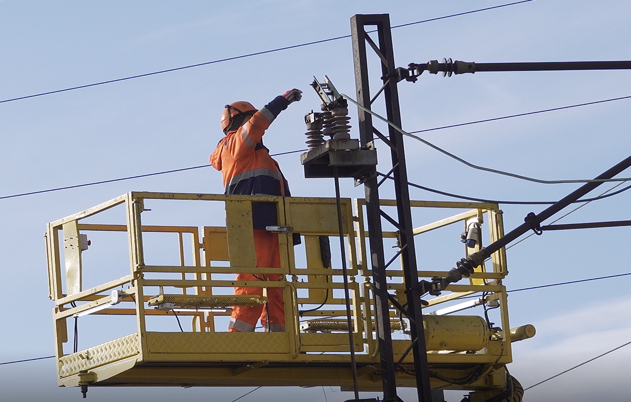 Zdjęcie obrazuje prace eksploatacyjne. Na zdjęciu widać żółtą platformę podnośnika zabezpieczoną barierkami i uniesioną obok szczytu słupa sieci trakcyjnej. Na platformie znajduje się pracownik kolei przeprowadzający prace eksploatacyjne na elementach sieci trakcyjnej na szczycie słupa.