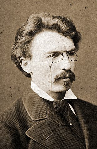 Zdjęcie portretowe młodego mężczyzny. Ma włosy zaczesane do góry, wąsy i binokle na nosie. Ubrany jest w elegancki ciemny garnitur z krawatem i białą koszulę.