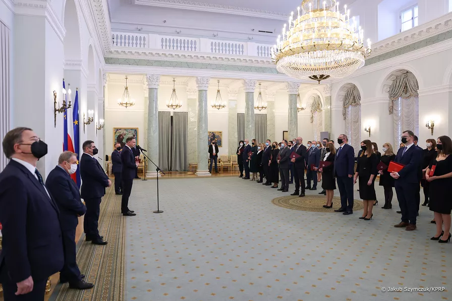 Zdjęcie przedstawia dużą salę pałacową z dwoma marmurowymi kolumnami. W sali stoją w dwóch rzędach ludzie w oficjalnych strojach. Część z nich trzyma w dłoniach czerwone teczki. Pod ścianą sali stoi przy mikrofonie Prezydent Andrzej Duda. Po jego bokach kilku mężczyzn w garniturach. Wszyscy zgromadzeni mają na twarzach maseczki.