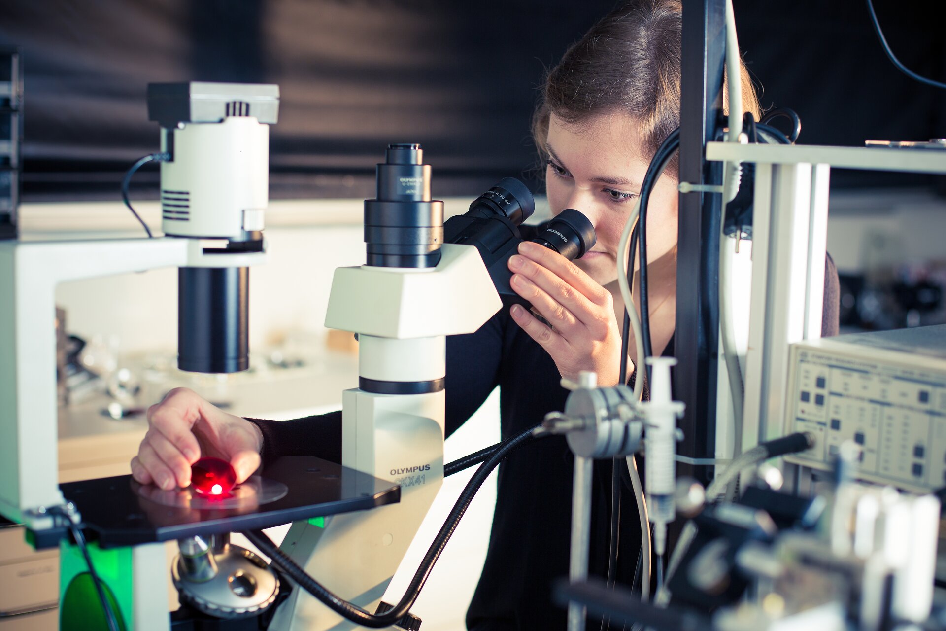 Na zdjęciu kobieta zerka do mikroskopu. Na szkiełku do obserwacji ma czerwoną kulkę. 