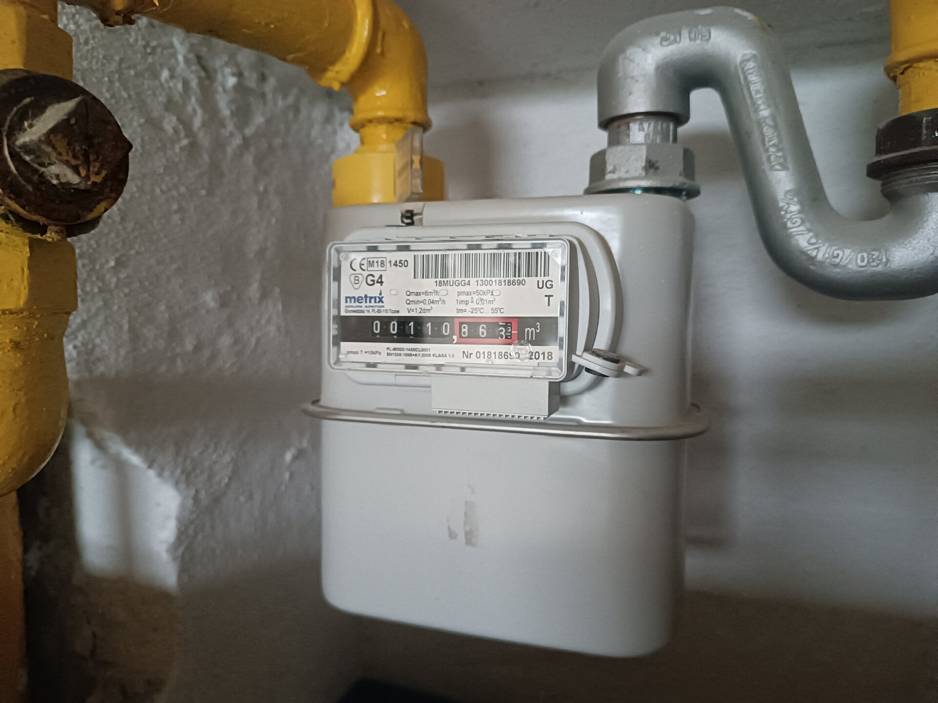 Na zdjęciu widoczny jest gazomierz miechowy zamontowany w gospodarstwie domowym. Gazomierz miechowy to urządzenie używane do pomiaru zużycia gazu w gospodarstwach domowych oraz innych budynkach. Przypomina niewielką metalową skrzynkę z wystającym okienkiem na przedniej stronie. Na okienku znajduje się kod kreskowy, informacje na temat urządzenia oraz licznik gazu. Do jego górnej części przymocowane są dwie rury.