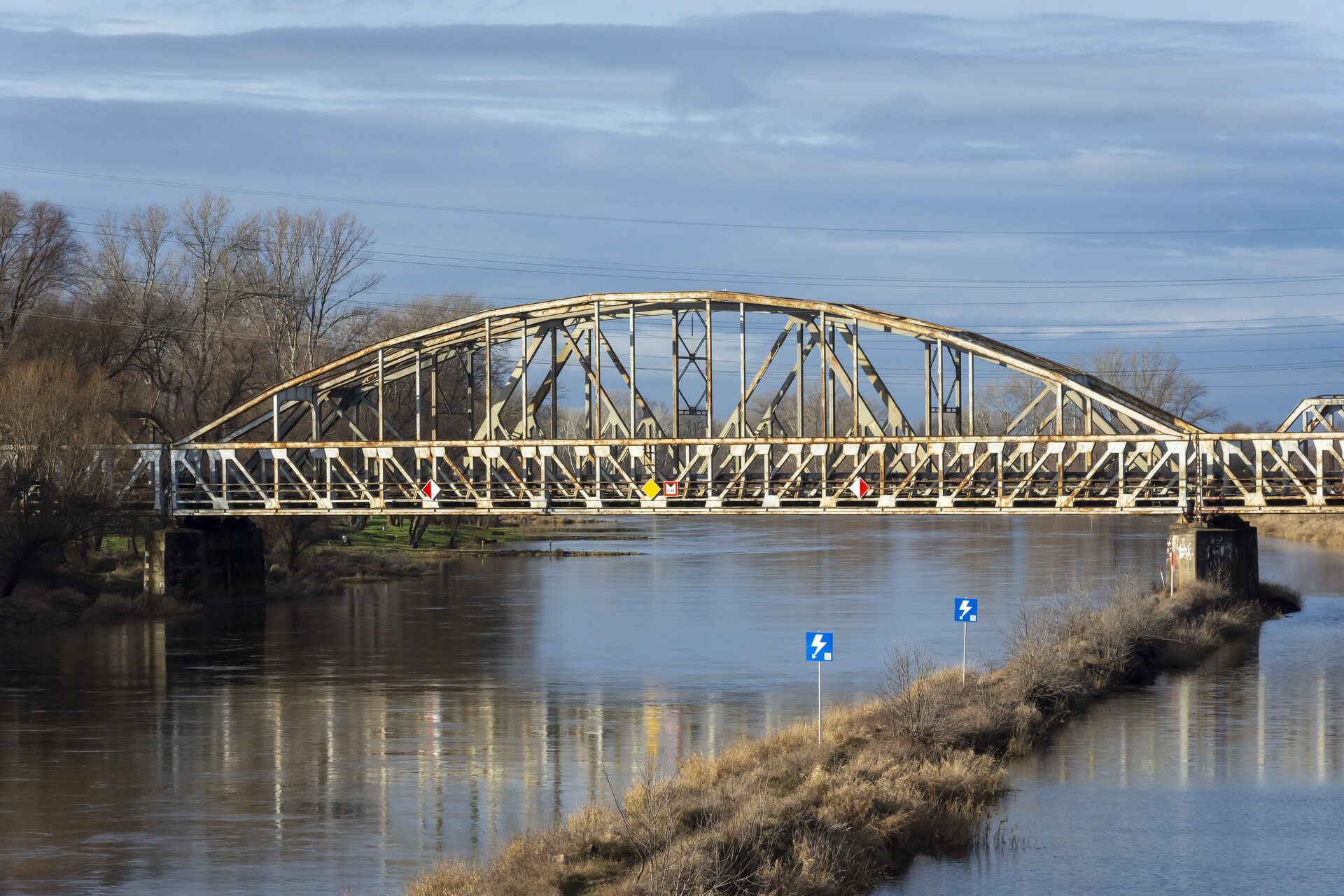 Ilustracja przedstawia most o konstrukcji łukowej, gdzie powierzchnia jezdna jest umiejscowiona u dołu. Konstrukcja mostu oparta jest na stalowej konstrukcji wspartej na żelbetowych podporach. Zarówno konstrukcja łuku, jak i kładka jezdna oraz bariery zostały pomalowane na biały kolor, z widocznymi śladami rdzy na niektórych elementach. Zdjęcie przedstawia most z boku, co pozwala dobrze zobaczyć żelbetowe podpory oraz łożyska między podporami a belkami.
