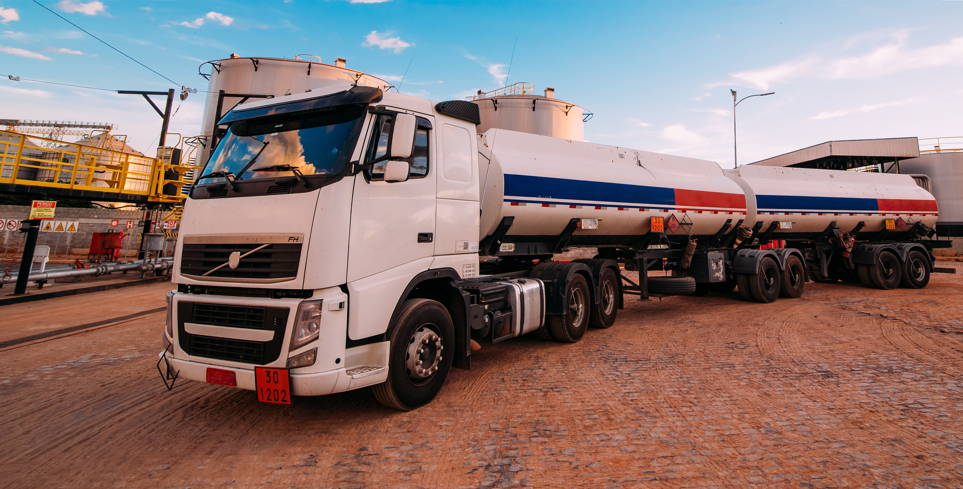 Ilustracja przedstawia pojazd do transportu drogowego ładunków niebezpiecznych. Pokazany jest samochód ciężarowy z długą naczepą z dwoma zbiornikami na ciecz w kształcie cylindrów leżących na podwoziu.