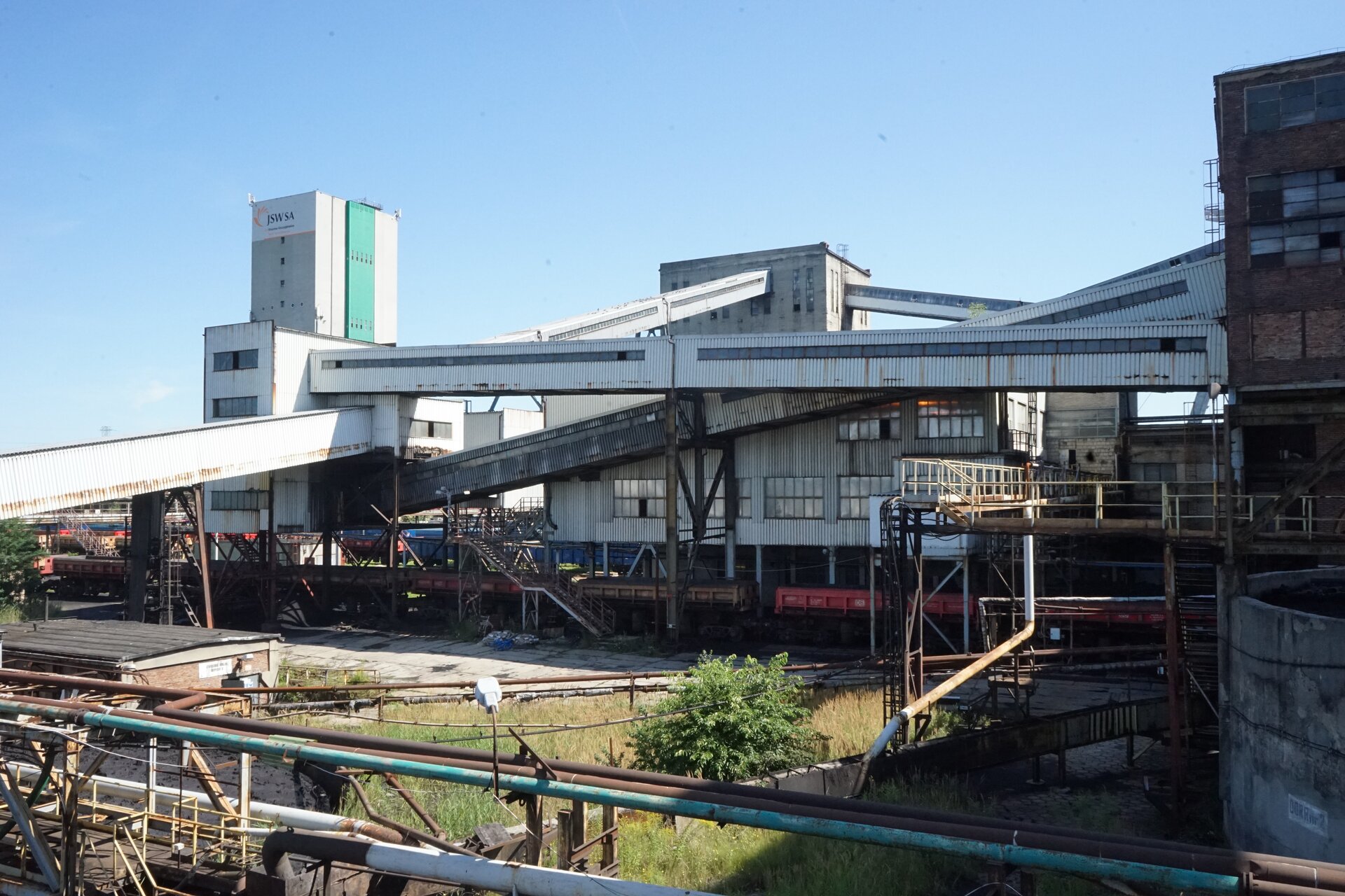 Na zdjęciu widoczny jest teren przemysłowy. W centralnej części zdjęcia znajdują się budynki przemysłowe o kształcie prostopadłościanu. W górnej części budynki są połączone mostami transportowymi. Mosty pod postacią łączników mają kształt prostopadłościanów. Mosty znajdują się na stalowych filarach. Mosty wykonane są z blachy falistej. W górnej części mostów wstawione są okna. Przy budynkach znajdują się tory kolejowe. Na torach widoczne są wagony transportowy. Przed torami widoczne są stalowe rury.Poniżej zdjęcia widoczny jest przycisk multimedialny z nagraniem audio. Treść nagrania jest zgodna z tytułem zdjęcia.