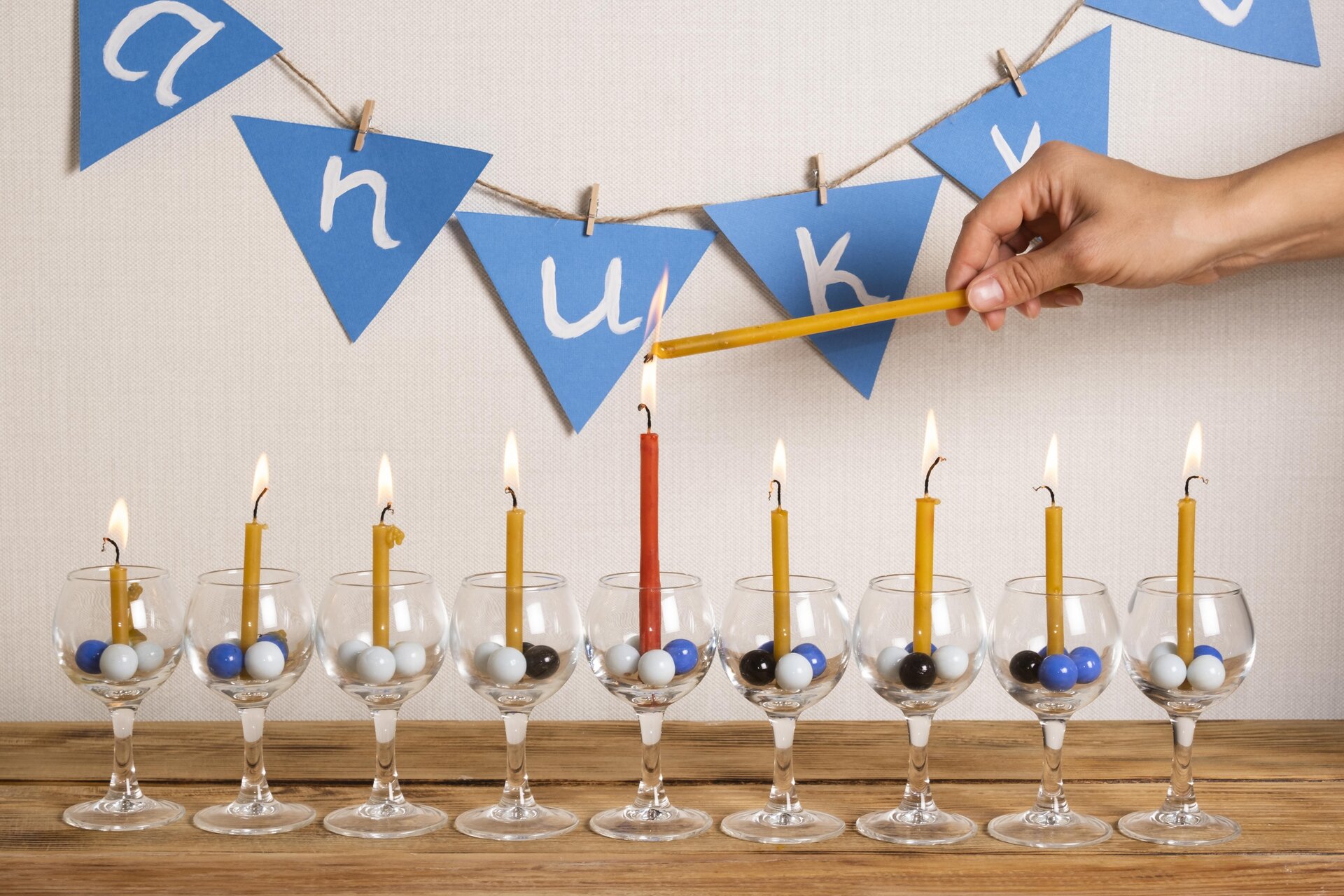 Zdjęcie przedstawia dziewięć kieliszków na wysokiej nóżce stojących na drewnianym blacie. W każdym kieliszku widoczne są małe szklane kulki w kolorach niebieskim, białym i czarnym, oraz cienkie palące się świece w kolorze żółtym. Świeca pośrodku ma kolor czerwony. Powyżej, po prawej stronie, widoczna jest dłoń trzymająca cienką świecę, której knot dotyka płomienia czerwonej świecy. Na ścianie widoczny jest łańcuch złożony z niebieskich chorągiewek, na których widoczne są pojedyncze białe litery. Od lewej: a, n, u, k, k, układające się w napis Hanukka , czyli Chanuka.