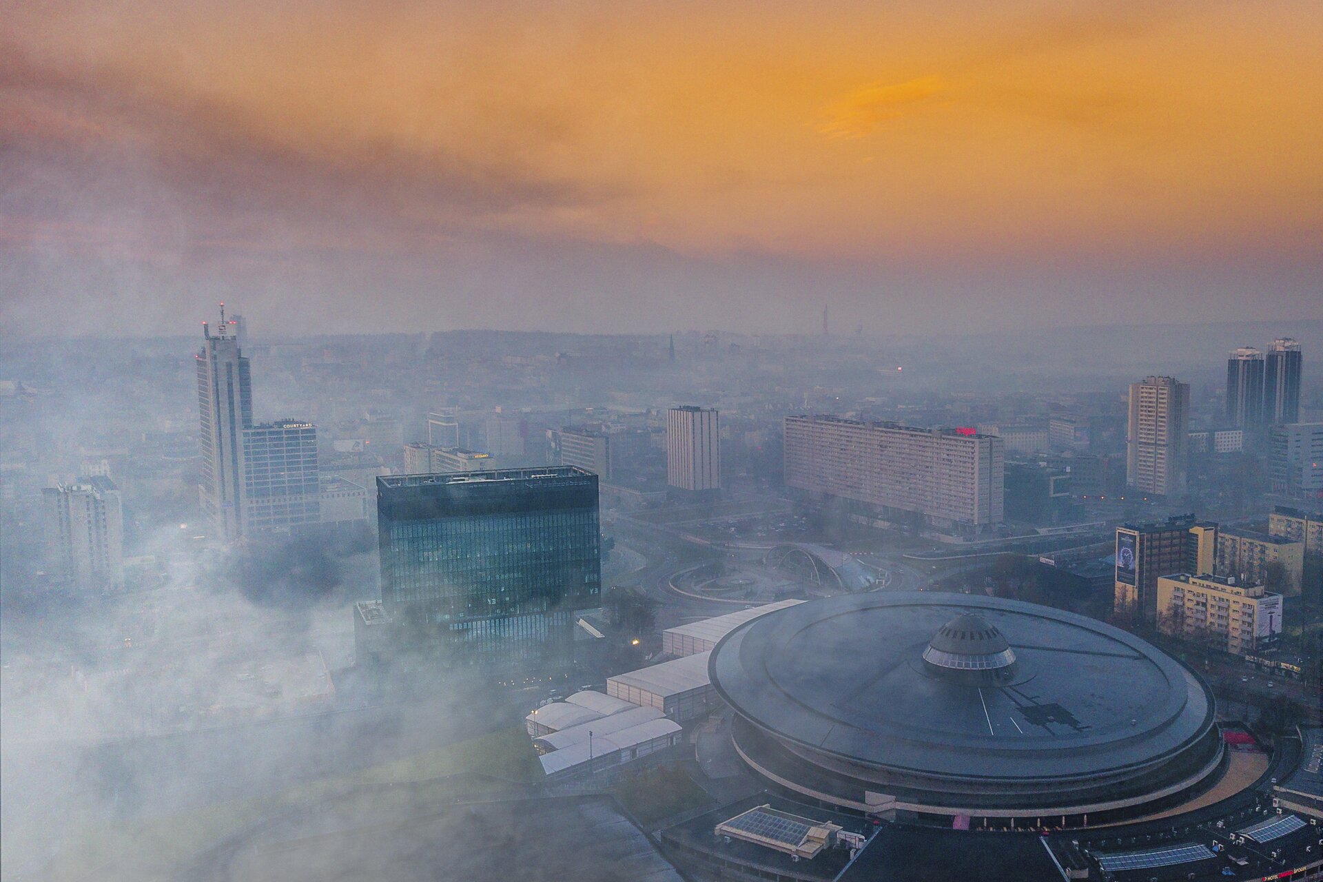 Fotografia lotnicza centrum Katowic. Na pierwszym planie katowicki Spodek, dookoła wieżowce, ulice i bloki. Nad miastem unosi się gęsty, szary smog ograniczający znacząco widoczność.