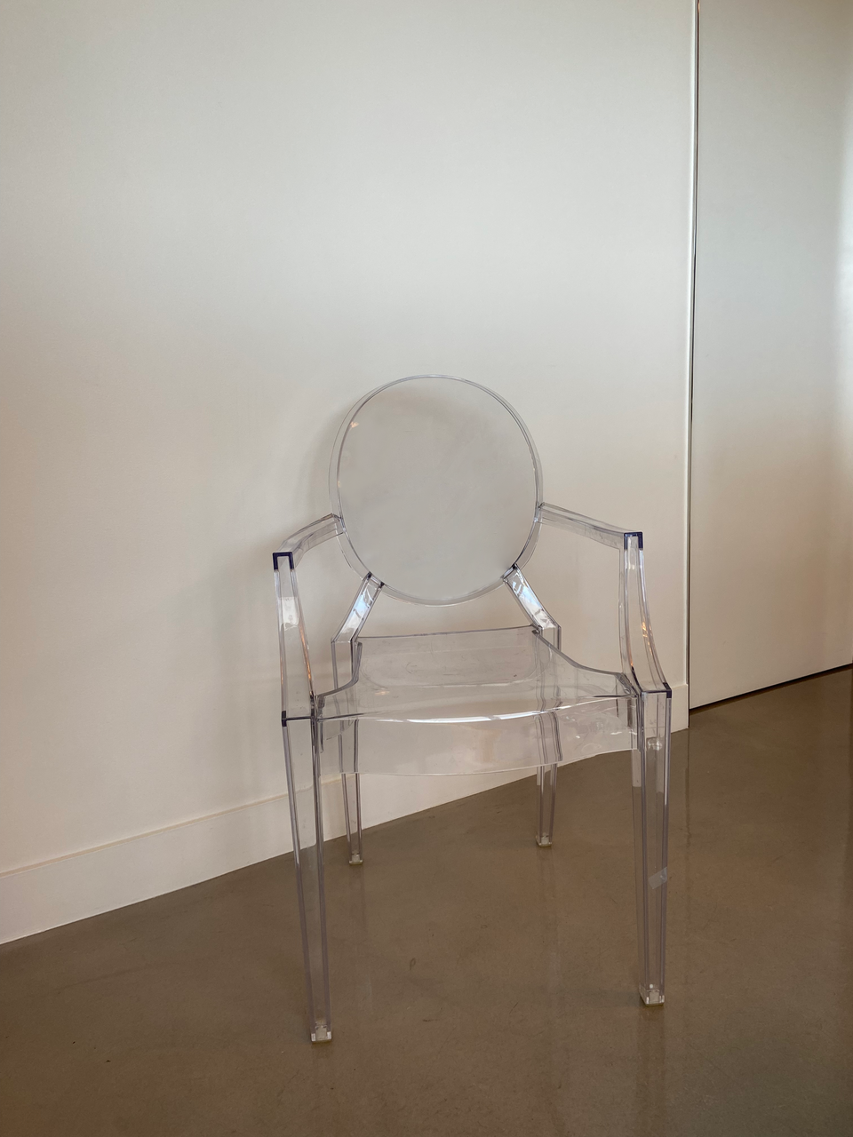 Zdjęcie przedstawia krzesło zrobione z przezroczystego materiału. Ma cztery nogi, podokietniki i owalne oparcie. Stoi na tle białej ściany i ciemnej podłogi.