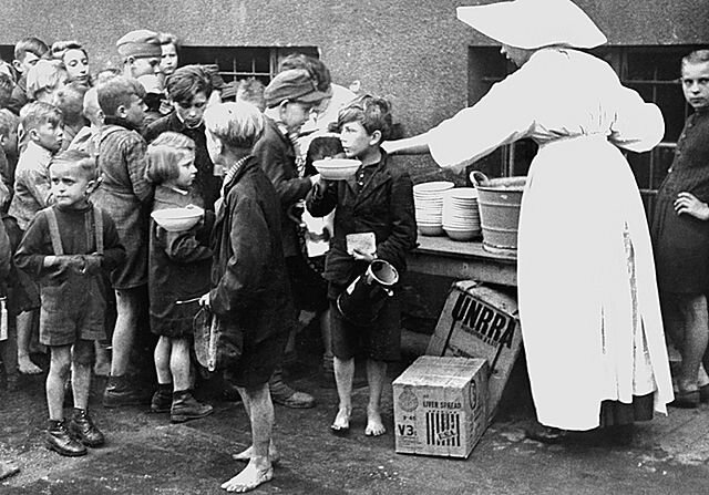 Zdjęcie przedstawia wydawanie posiłku dzieciom przez zakonnicę. Dzieci stoją w kolejce. Są skromnie ubrane, część z nich jest bosa, w za dużych lub za małych ubraniach.
