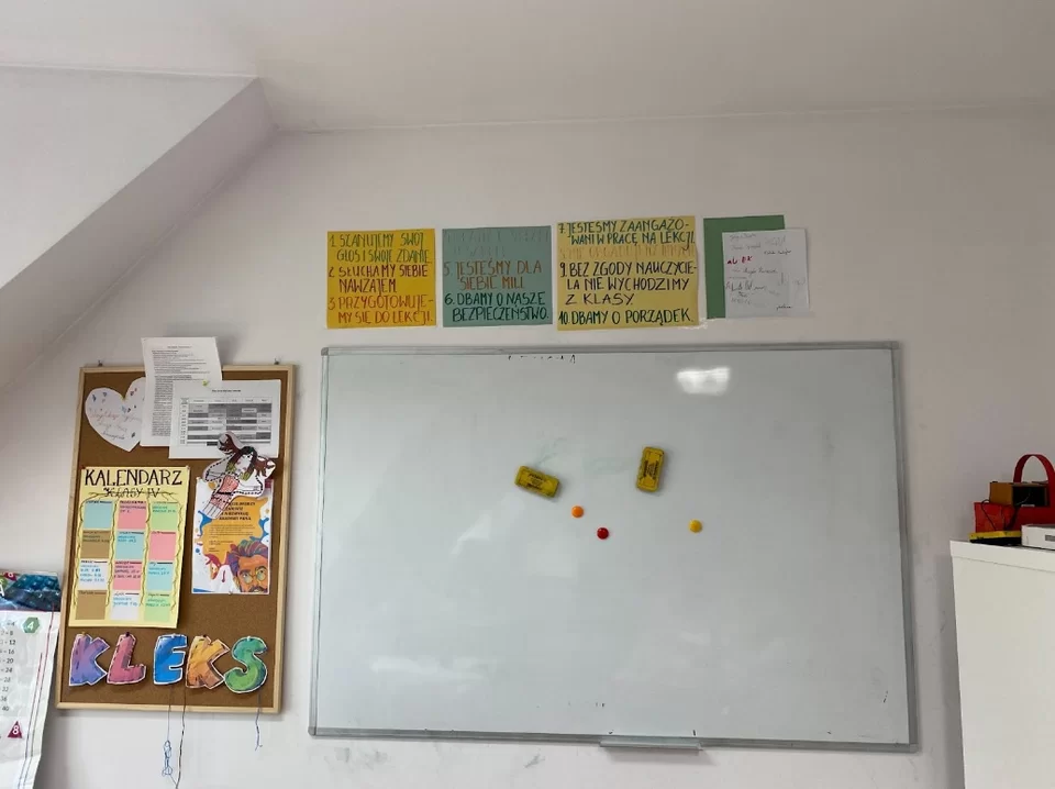 Na ścianie wisi tablica magnetyczna, obok niej z lewej strony wisi tablica korkowa z ważnymi informacjami; nad tablicą magnetyczną przyklejono kontrakt spisany kolorowymi flamastrami na dużych kartkach w różnych kolorach.