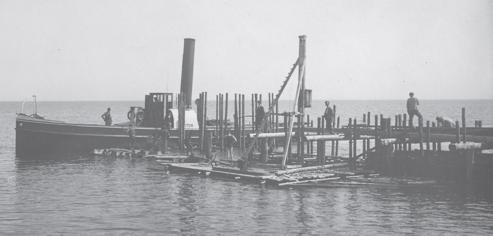 Zdjęcie przedstawia długą łódź na morzu w otoczeniu rusztowań. Z wody wystają długie belki. Łączone są one z belkami układanymi poziomo. Po rusztowaniach chodzą mężczyźni. Na łodzi stoi człowiek, który przygląda się pracy reszty osób. Fotografia jest czarno‑biała.